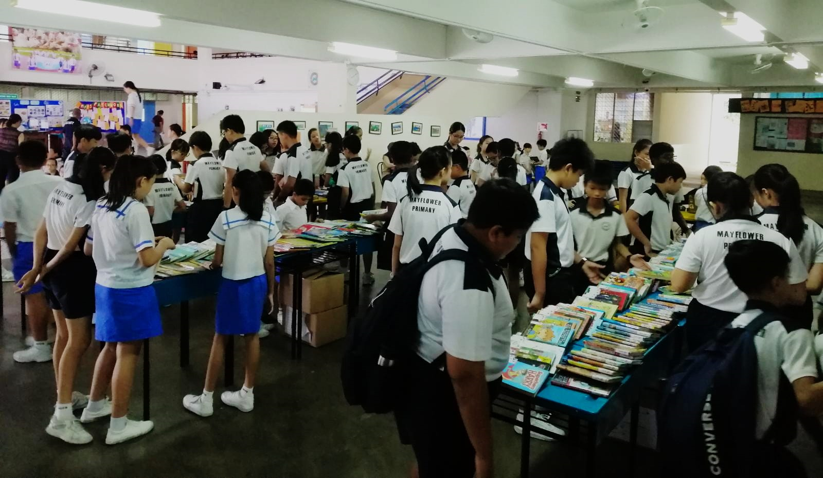 Mayflower Primary students in uniforms browse tables filled with books in a school hall.