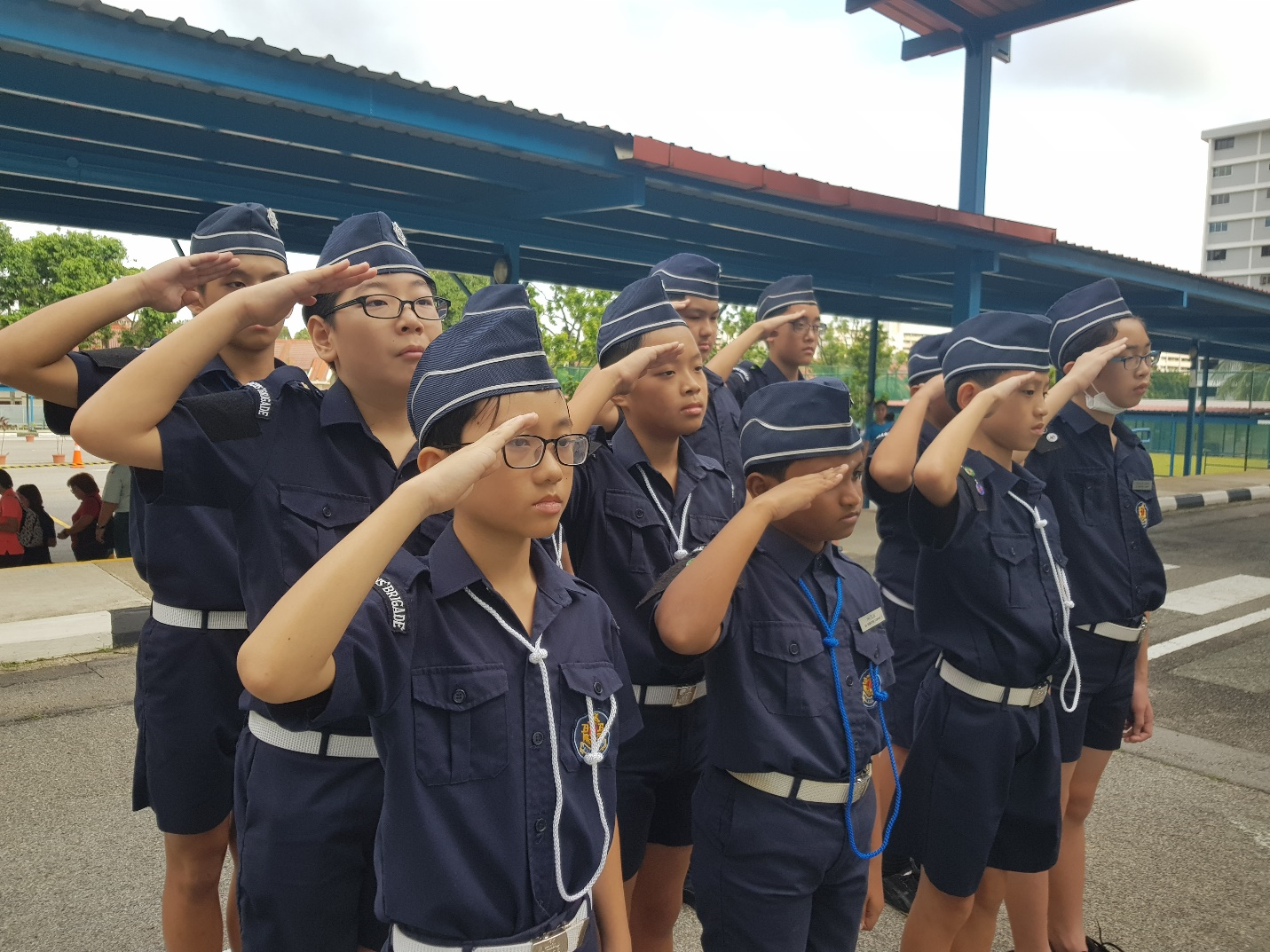 A group of children in navy blue uniforms and caps saluting in formation outdoors near a covered structure.