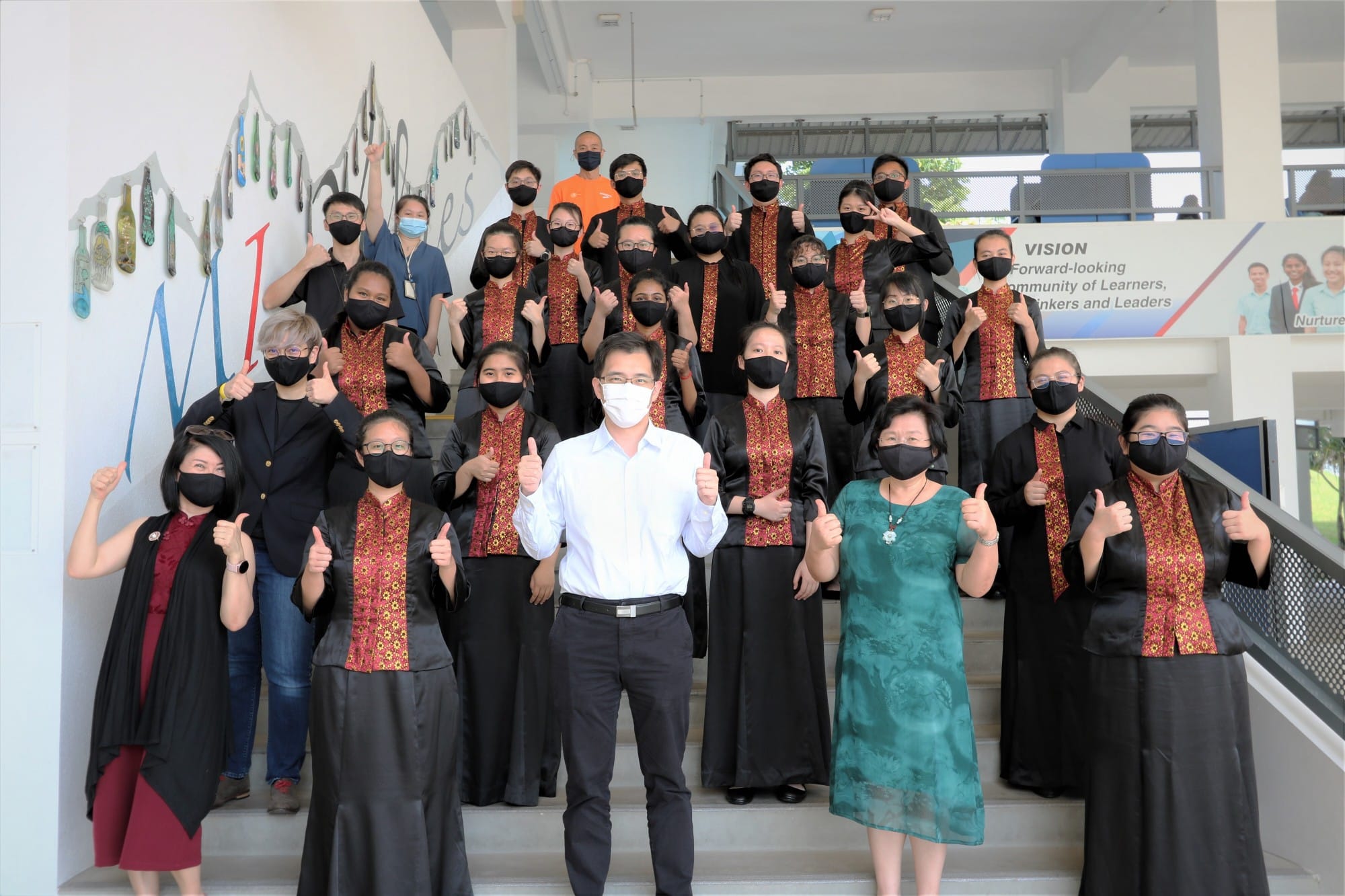 A large group wearing masks and matching black and red outfits pose on school stairs giving thumbs up.