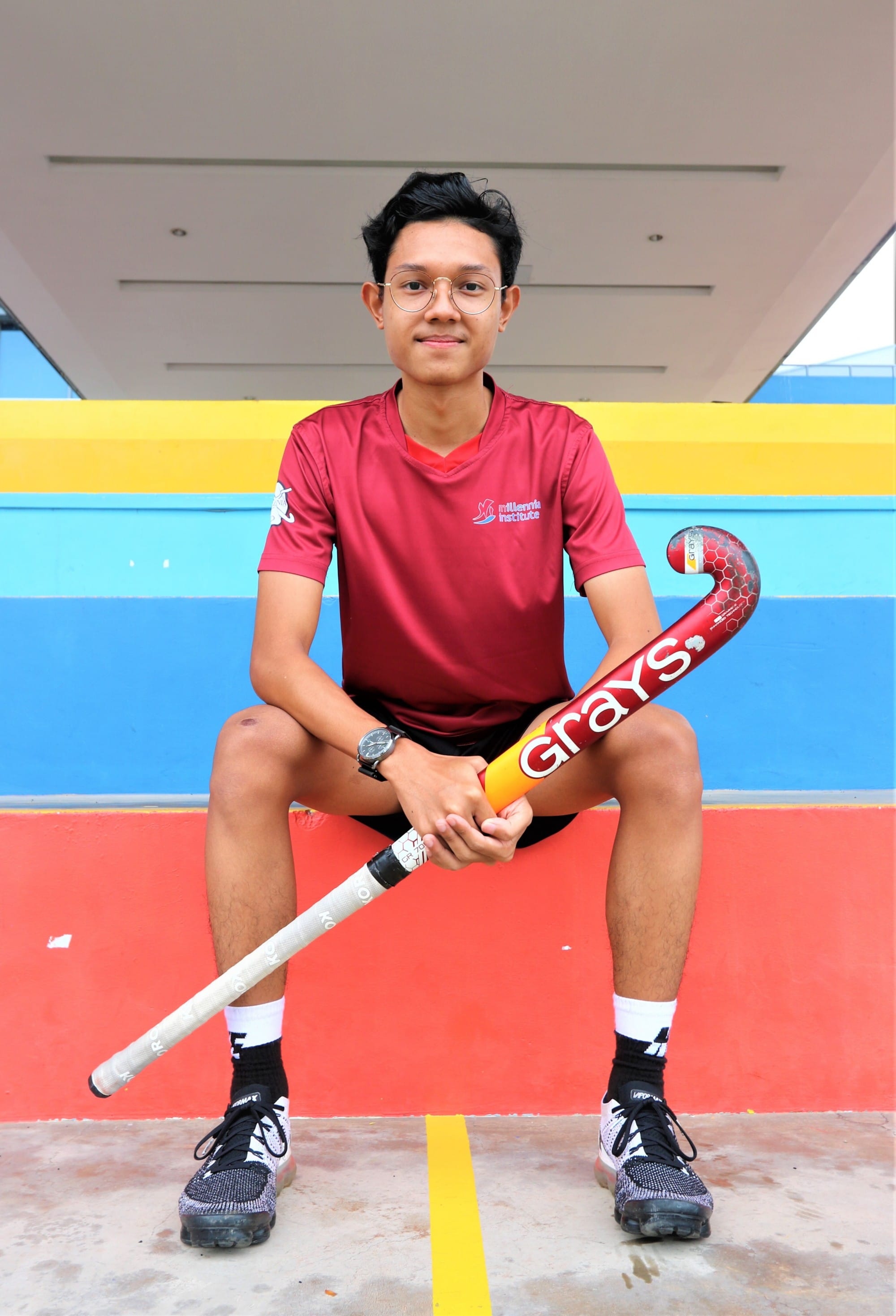 Young man in a red jersey sitting on bleachers, holding a Grays field hockey stick, wearing glasses.