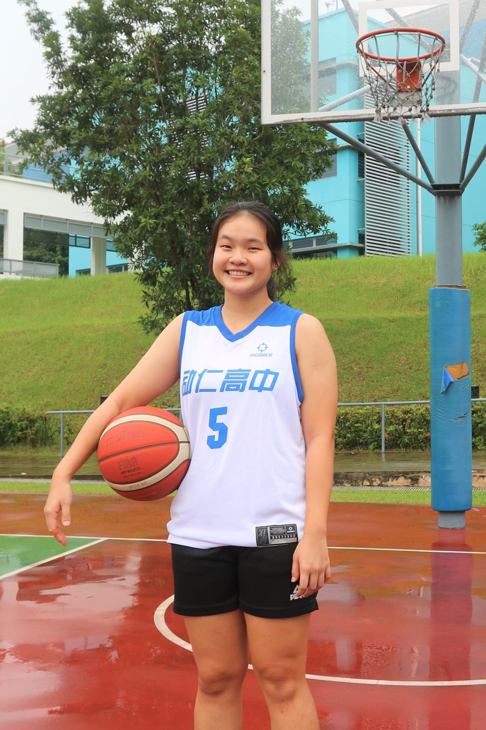 Young girl in jersey number 5 smiling on a wet outdoor basketball court, holding a basketball beside a hoop.