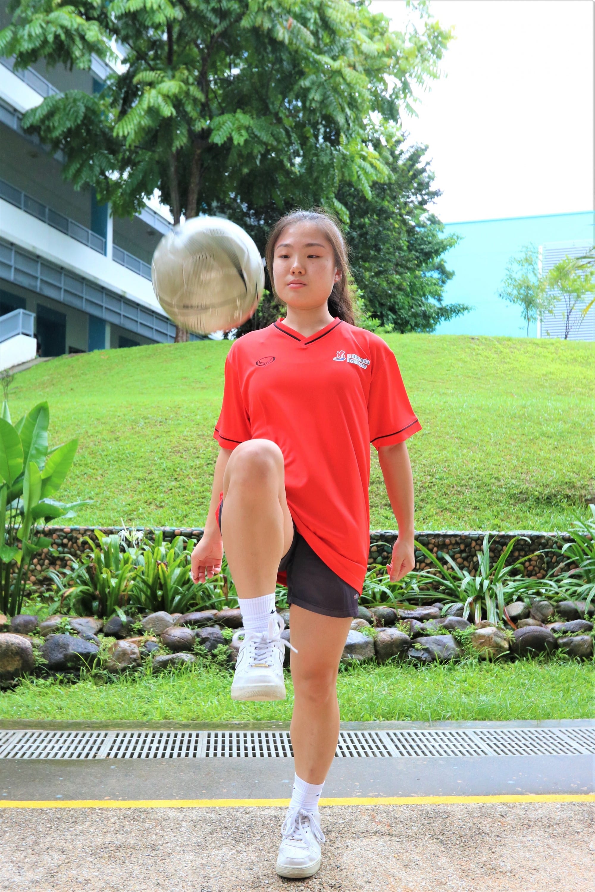 Young girl in a red jersey kicking a silver ball outdoors near a grassy hill and building.