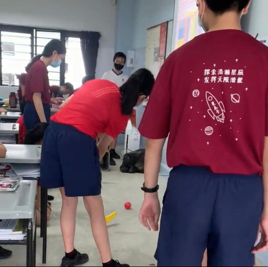 Students in red shirts and navy shorts stand in a classroom; one bends forward near a small red ball on the floor.