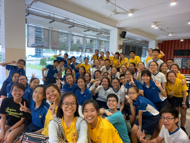 Large group of students in blue and yellow uniforms smiling and making peace signs in a school hall.