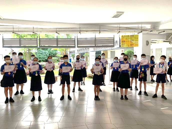 Students in blue uniforms and masks stand in rows outdoors, each holding a certificate or award.