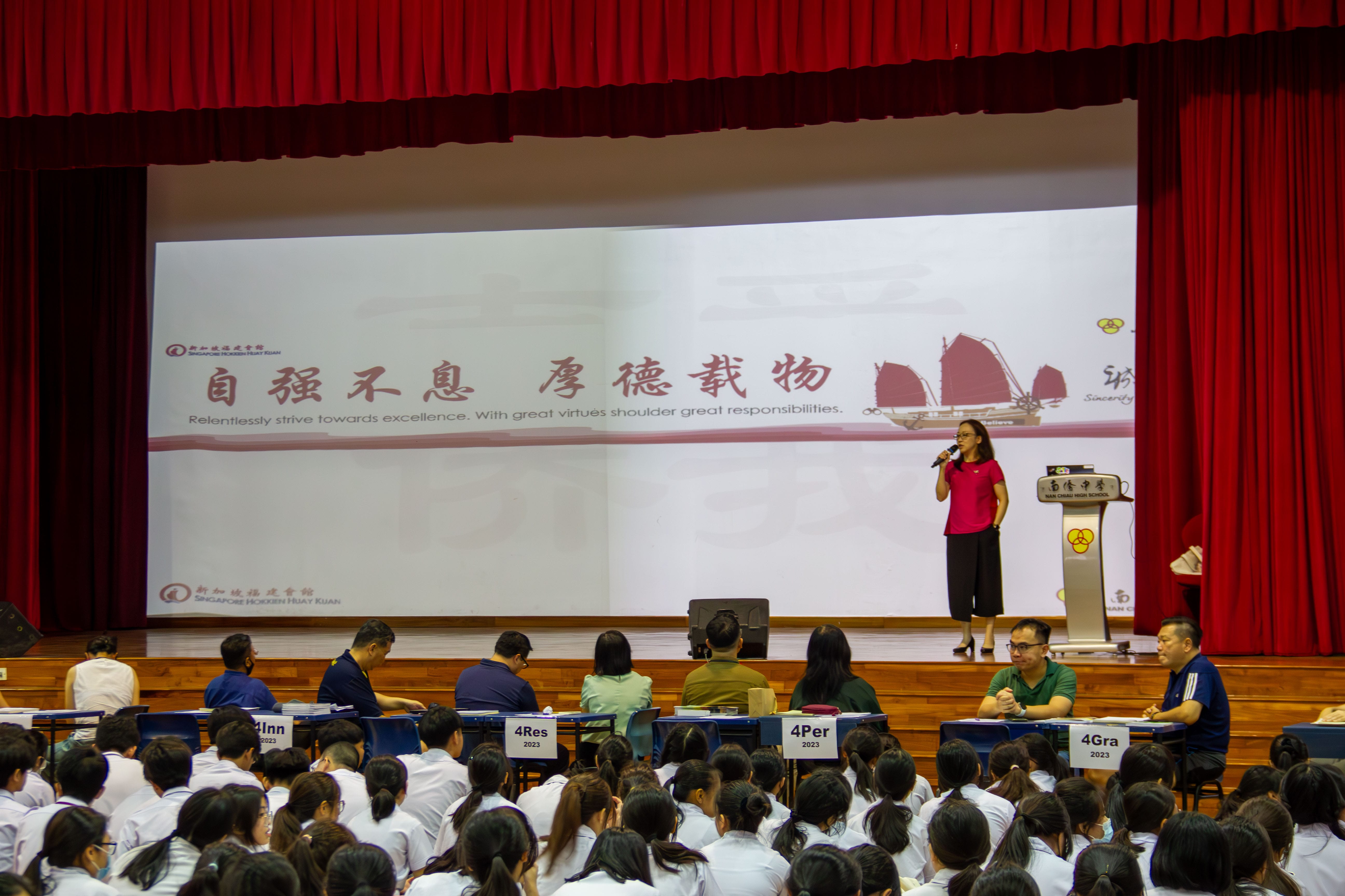 A woman in a red top speaks on stage at Nan Chiau High School while students in white uniforms sit in the audience.