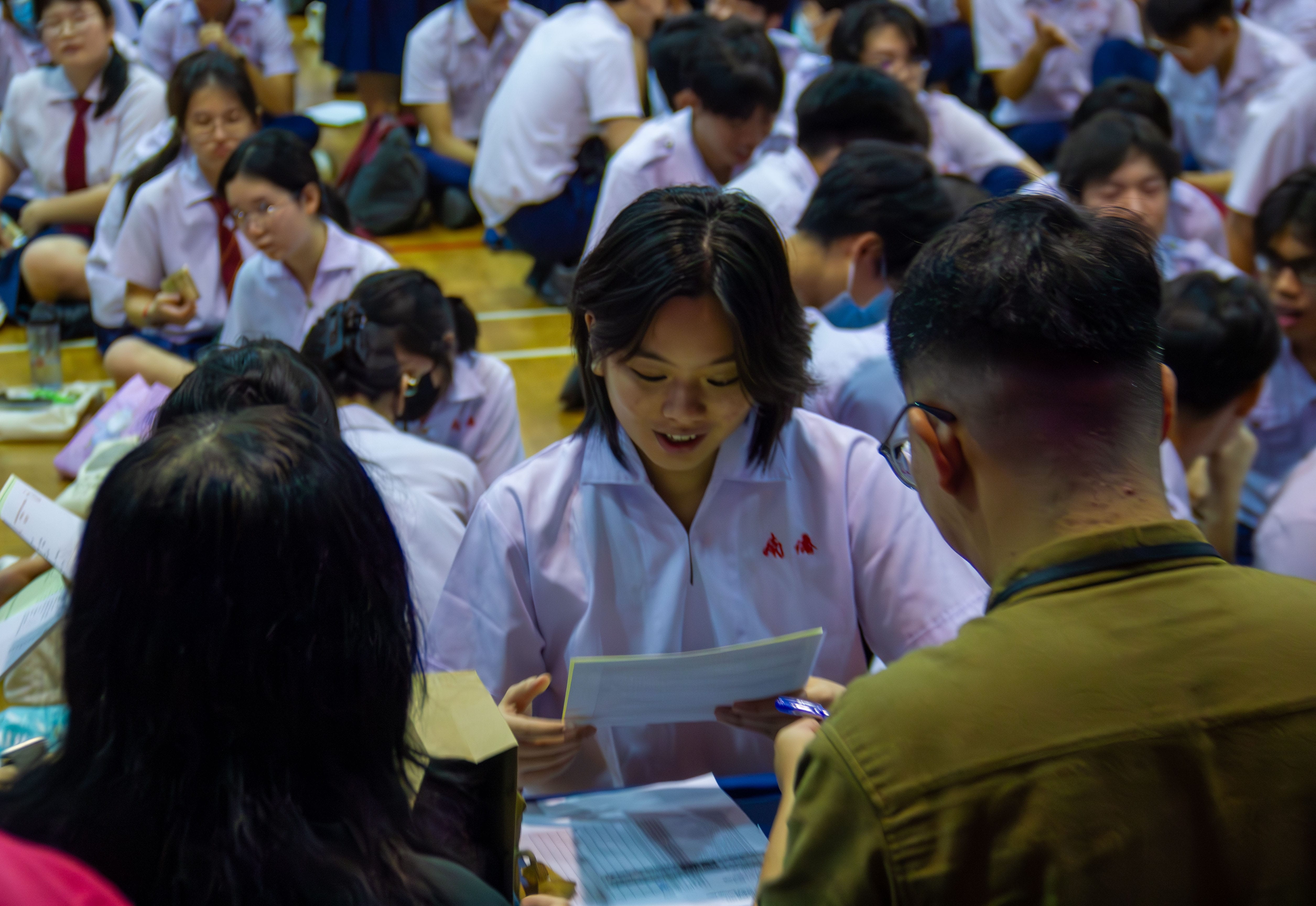 A girl in a white school uniform holds papers and speaks to two people in a crowded school hall filled with students.