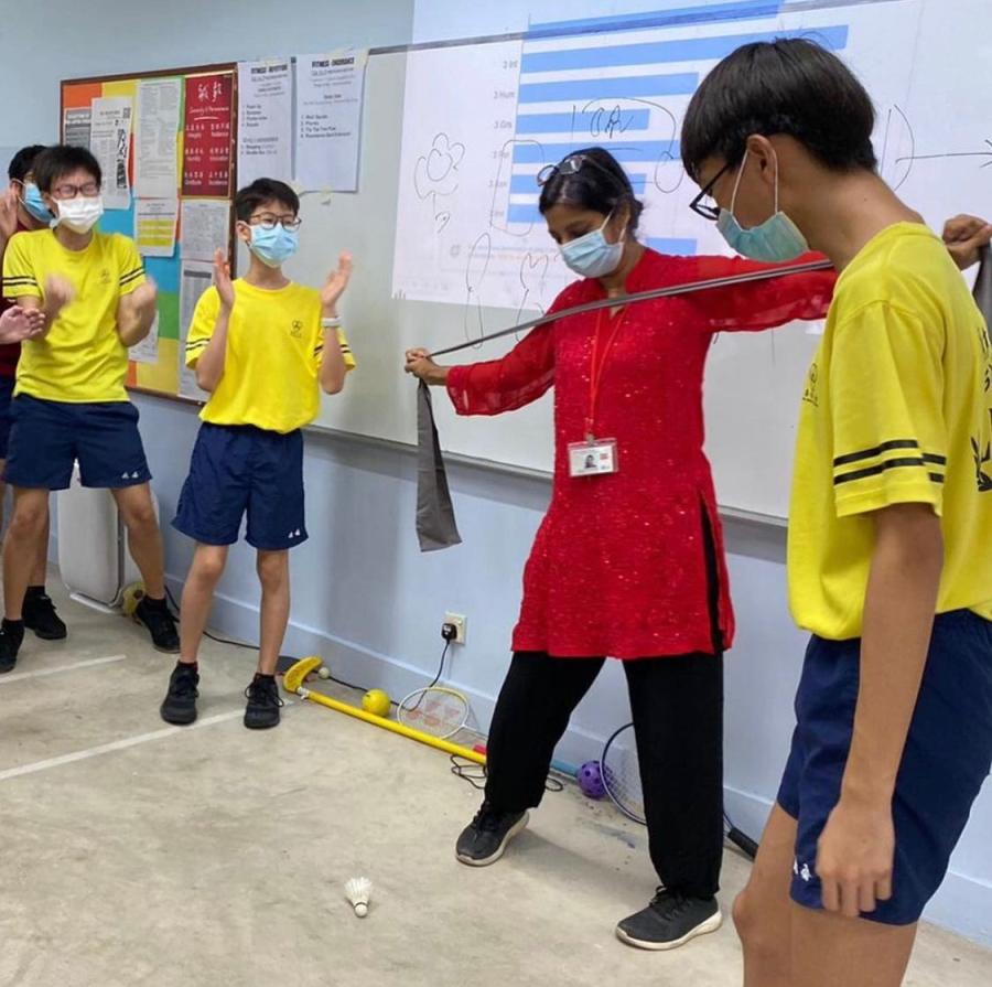 A masked teacher in red holds a resistance band with a student while others in yellow uniforms watch in a classroom.
