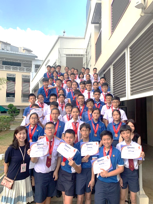 A large group of uniformed students stand on outdoor school steps, several holding certificates and wearing red medal ribbons.