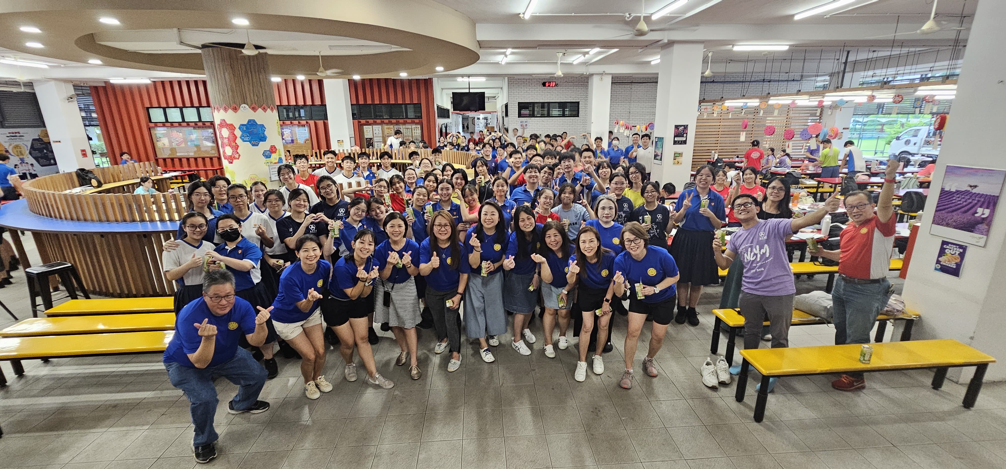 Large group of people, many in blue shirts, posing and smiling in a bright indoor canteen with yellow benches.