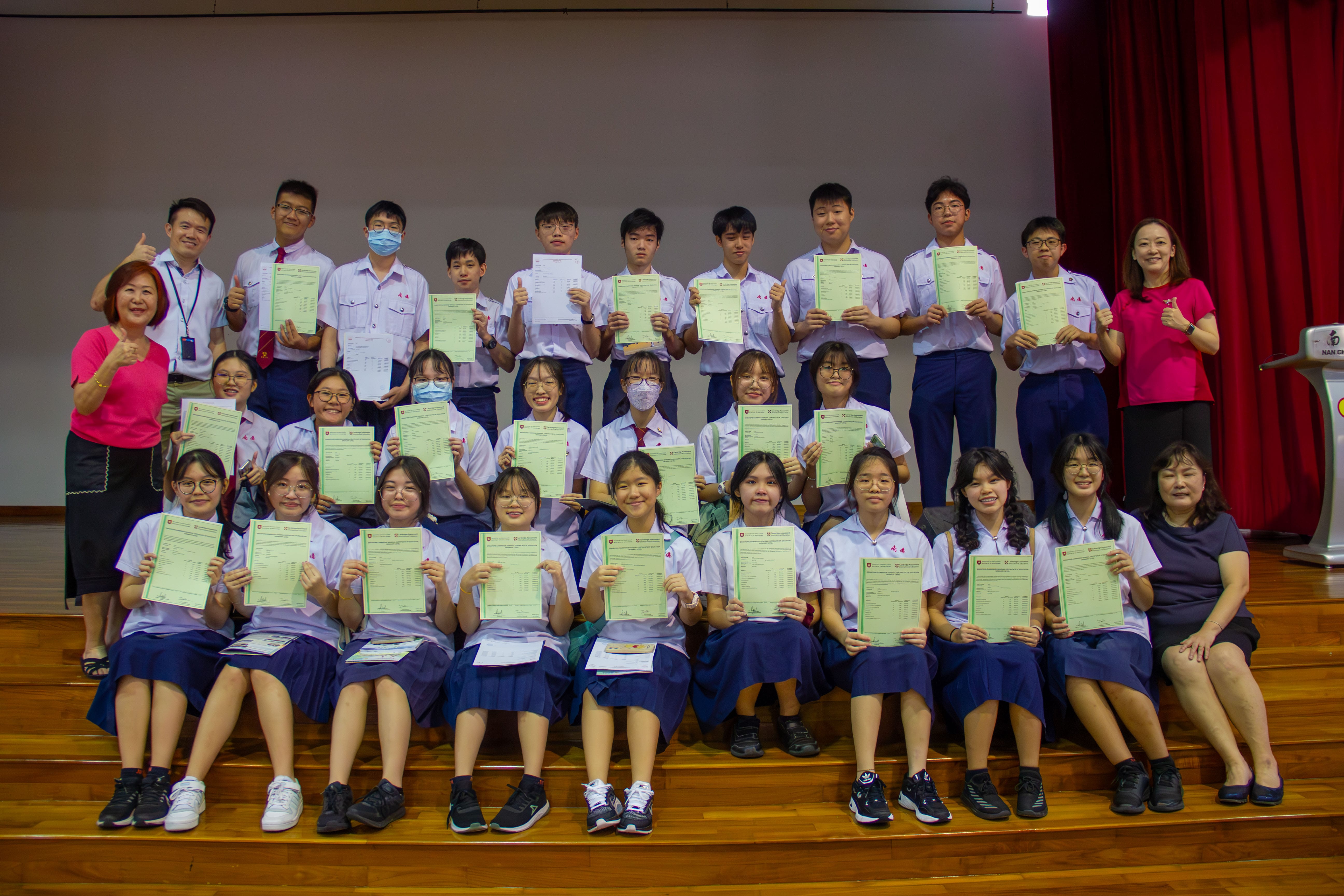 A large group of school students in uniforms hold up green certificates on a stage, posed with two adult teachers.
