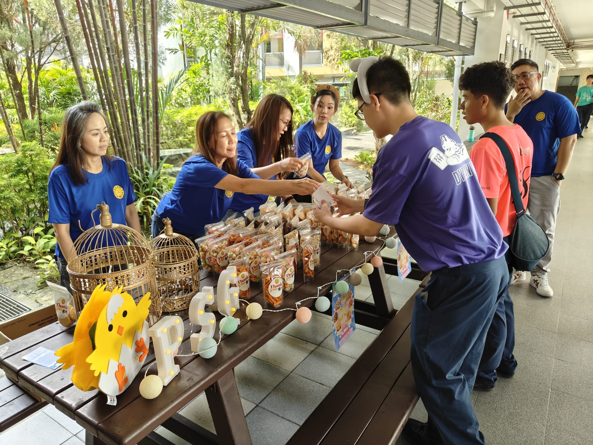 Three women in blue shirts hand out packaged snacks to students at an outdoor table decorated with birdcages and lights.