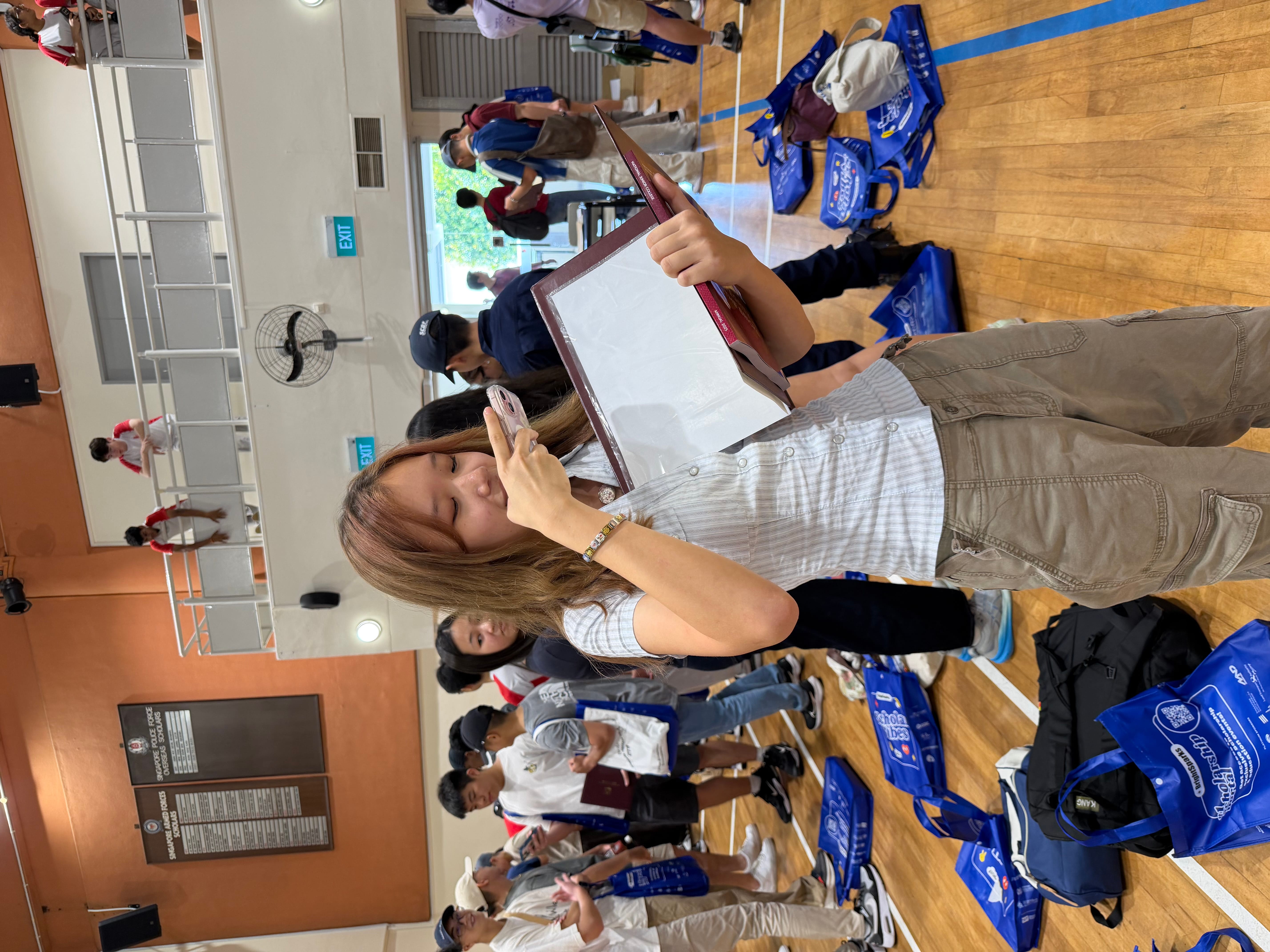 Woman holds a clipboard and phone in a gym, with people and blue bags scattered on the floor behind her.