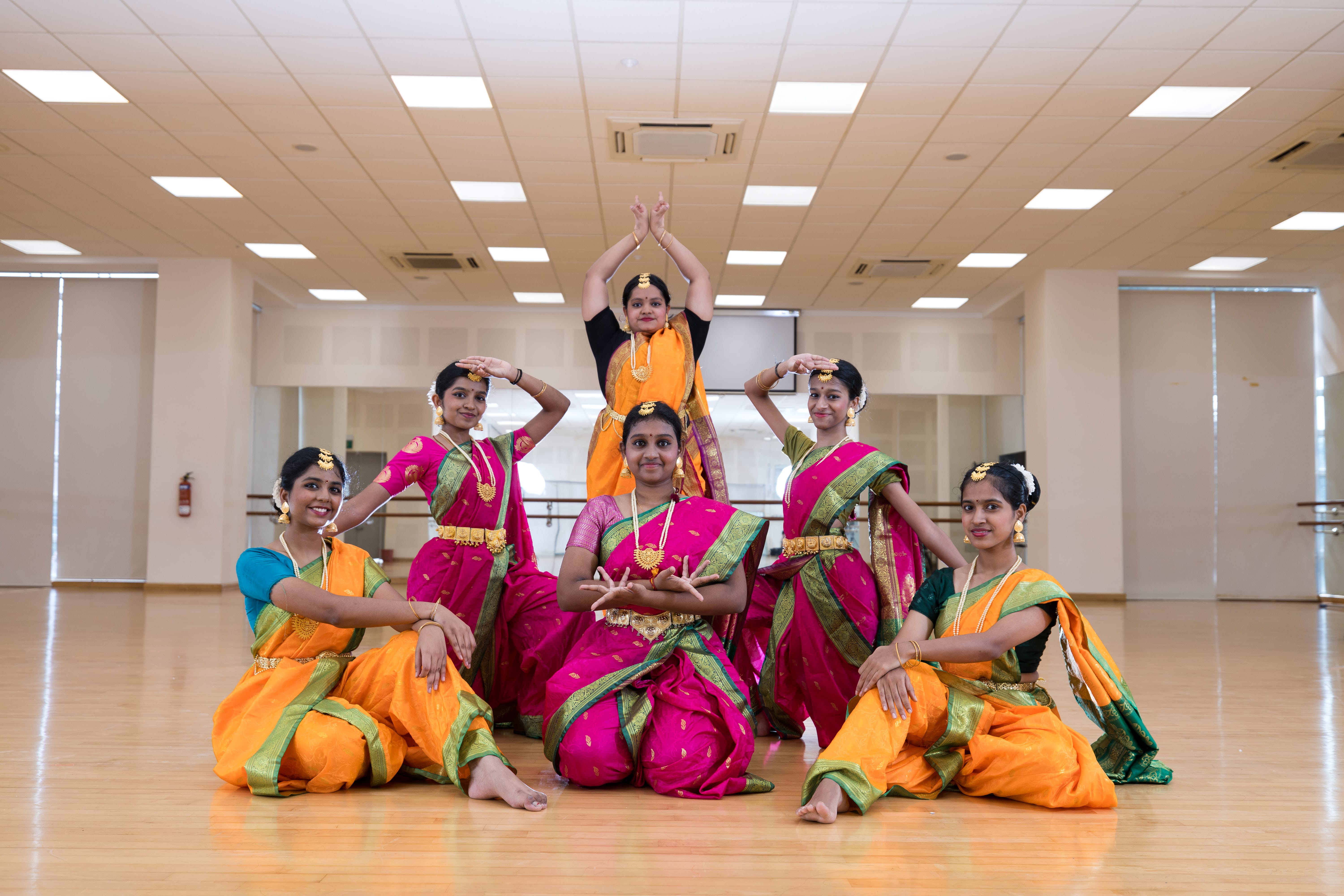 Six women in colorful saris pose in a dance studio, seated and standing with arms raised.