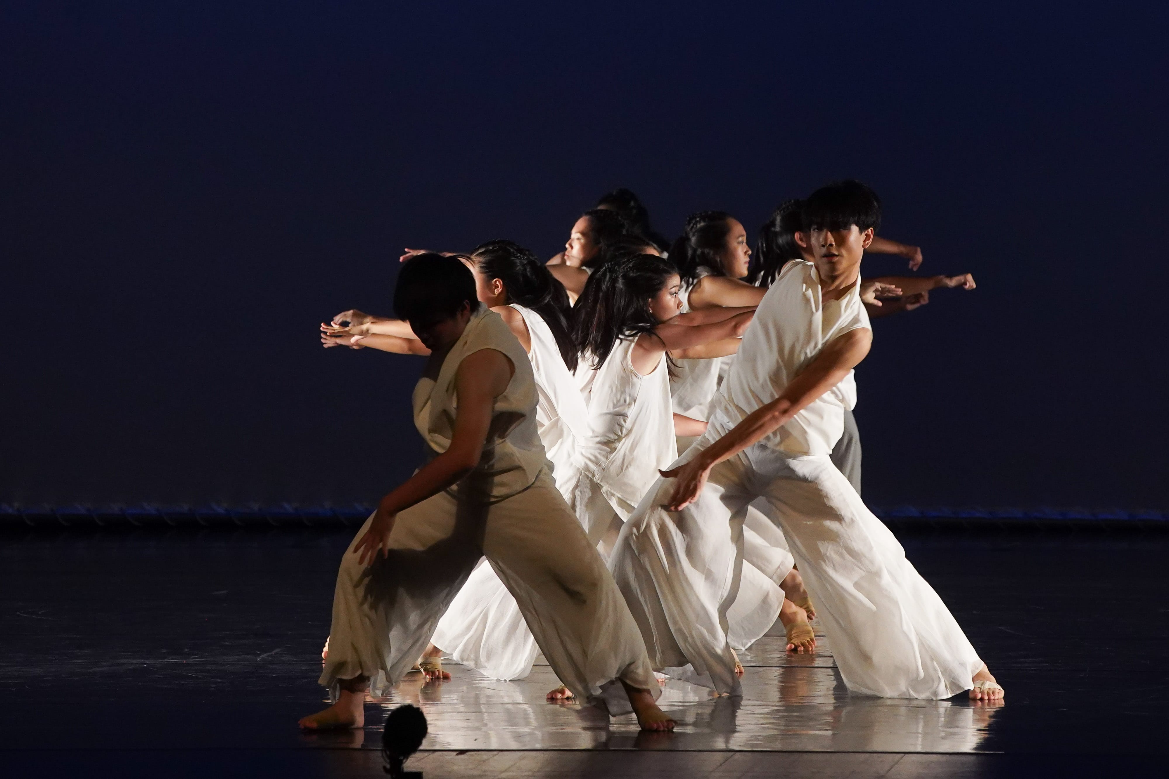 Several dancers in white flowing garments perform a synchronized contemporary dance on a dark stage.