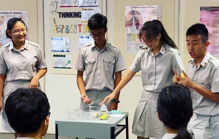 Four students in grey uniforms conduct a classroom science experiment at a table with a clear container.