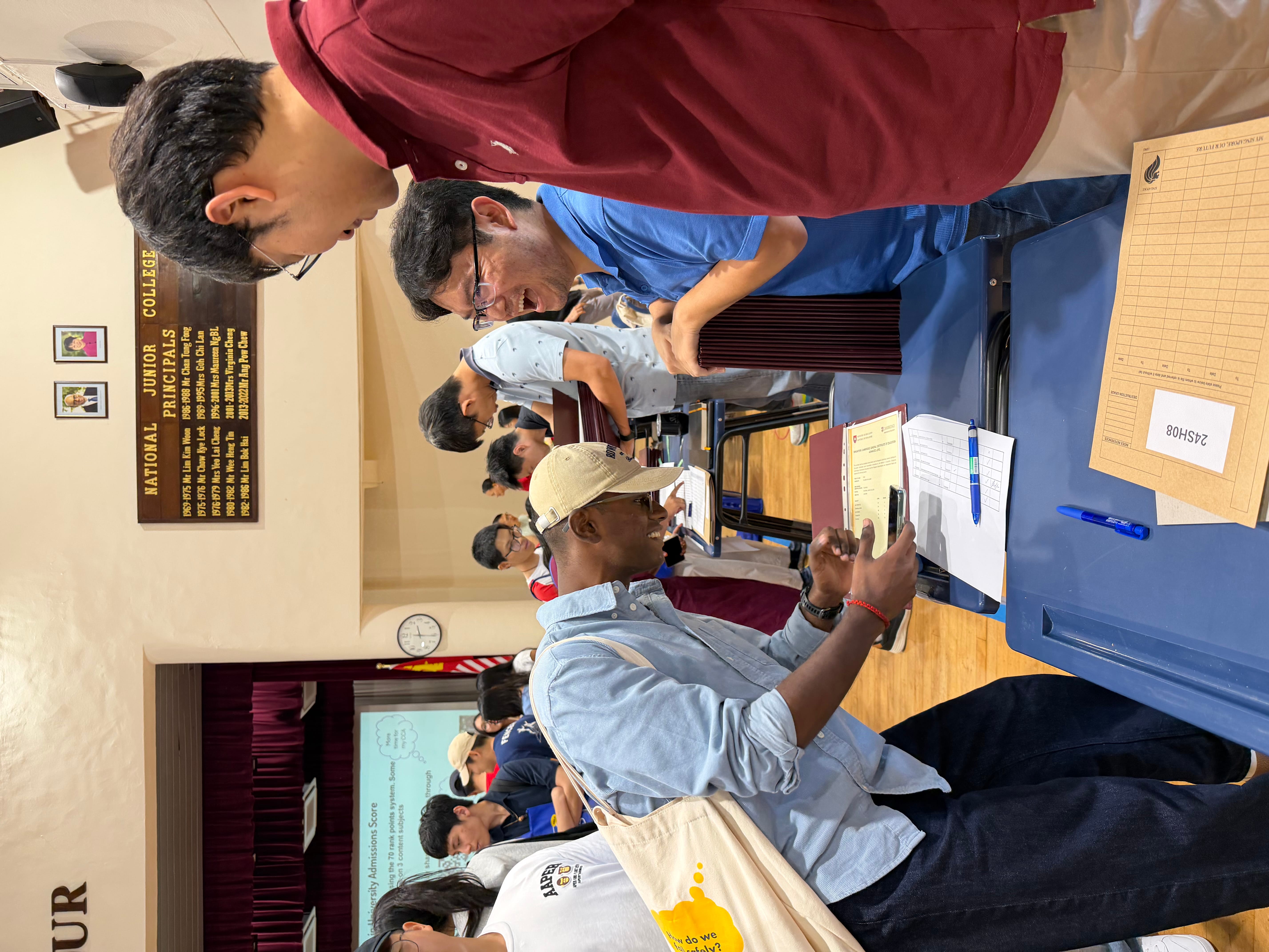People at tables in a hall; a man in a cap checks his phone beside clipboards and pens.