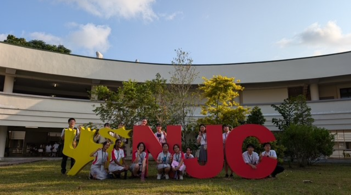 Students pose in front of large red NJC letters and a yellow mascot sign outside a curved school building.