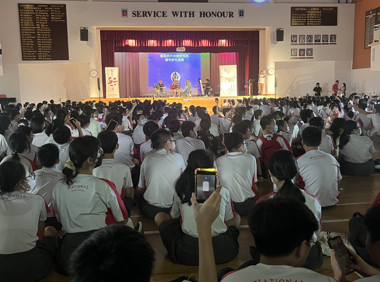 National Junior College students in uniforms sit on a hall floor watching a stage performance under a "Service With Honour" sign.