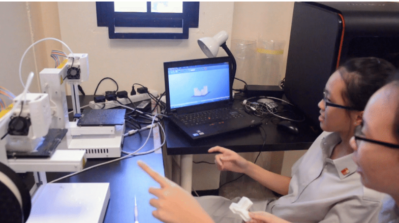 Two students wearing glasses sit at a lab desk with a laptop and 3D printer, pointing at the screen.
