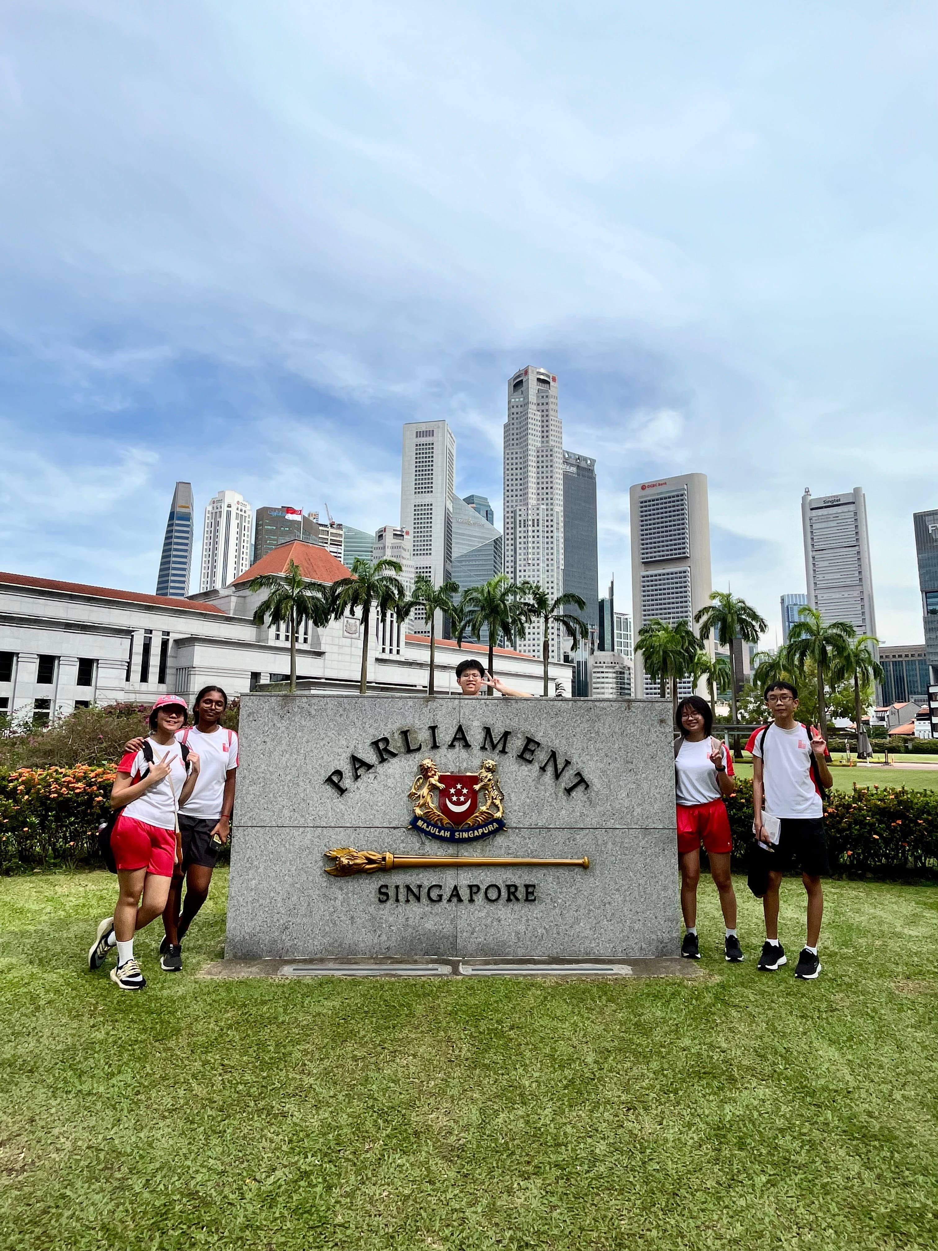 Four students in white and red pose beside the Parliament Singapore stone sign with city skyscrapers behind.