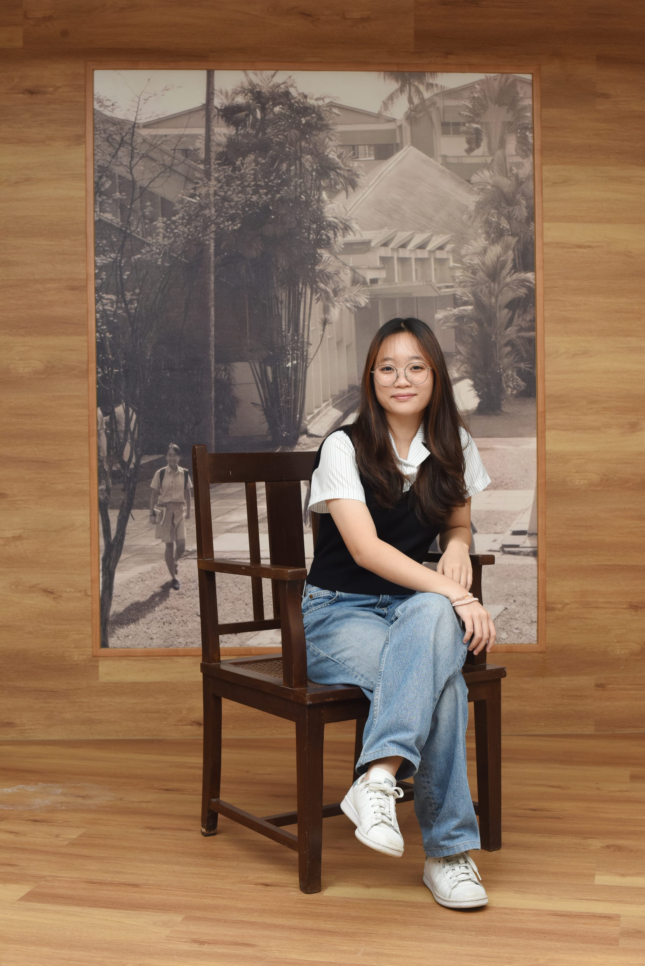 Young woman with glasses and long dark hair sits on a wooden chair before a vintage black-and-white photo mural.