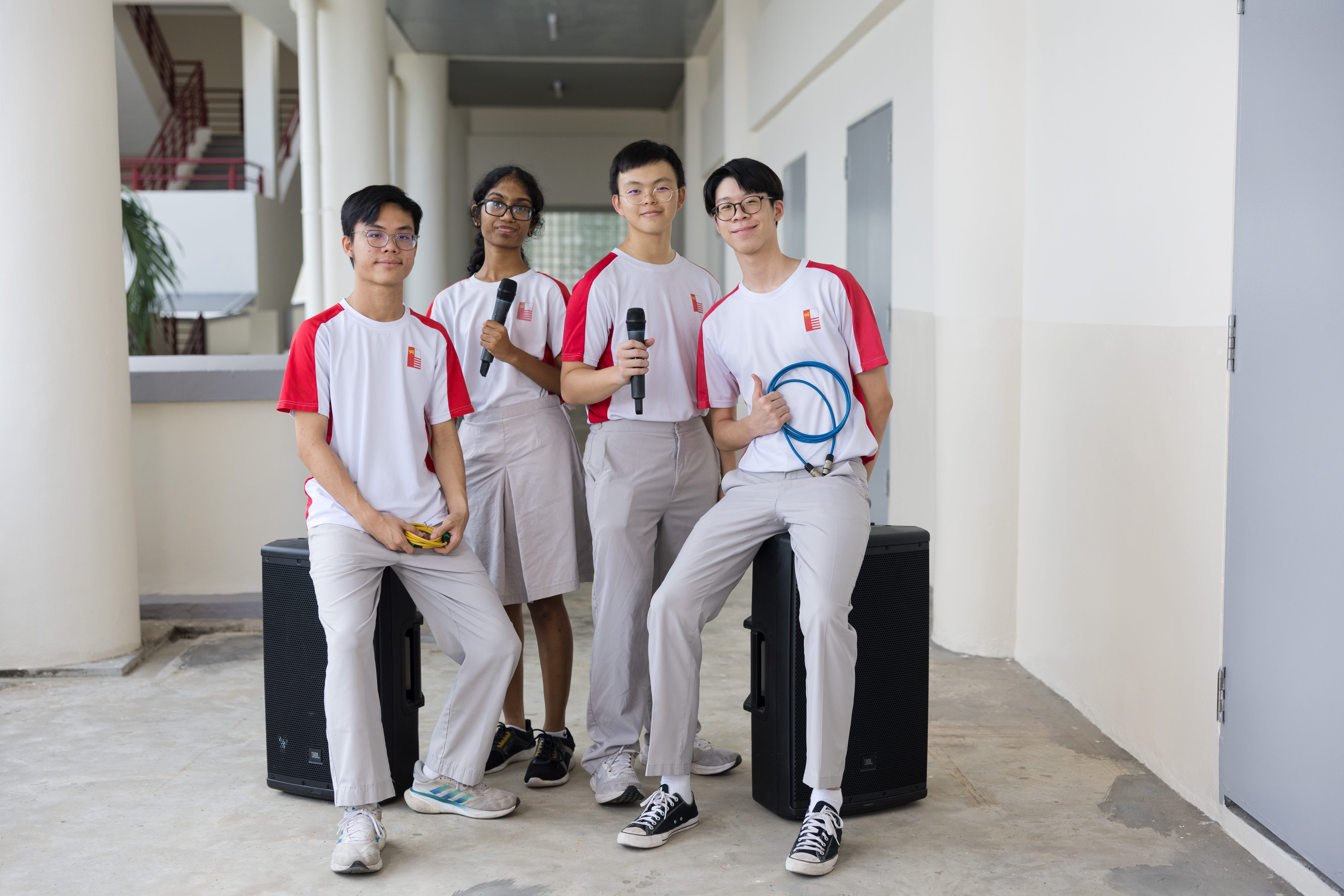 Four students in white and red uniforms pose in a hallway with microphones and two black speakers.