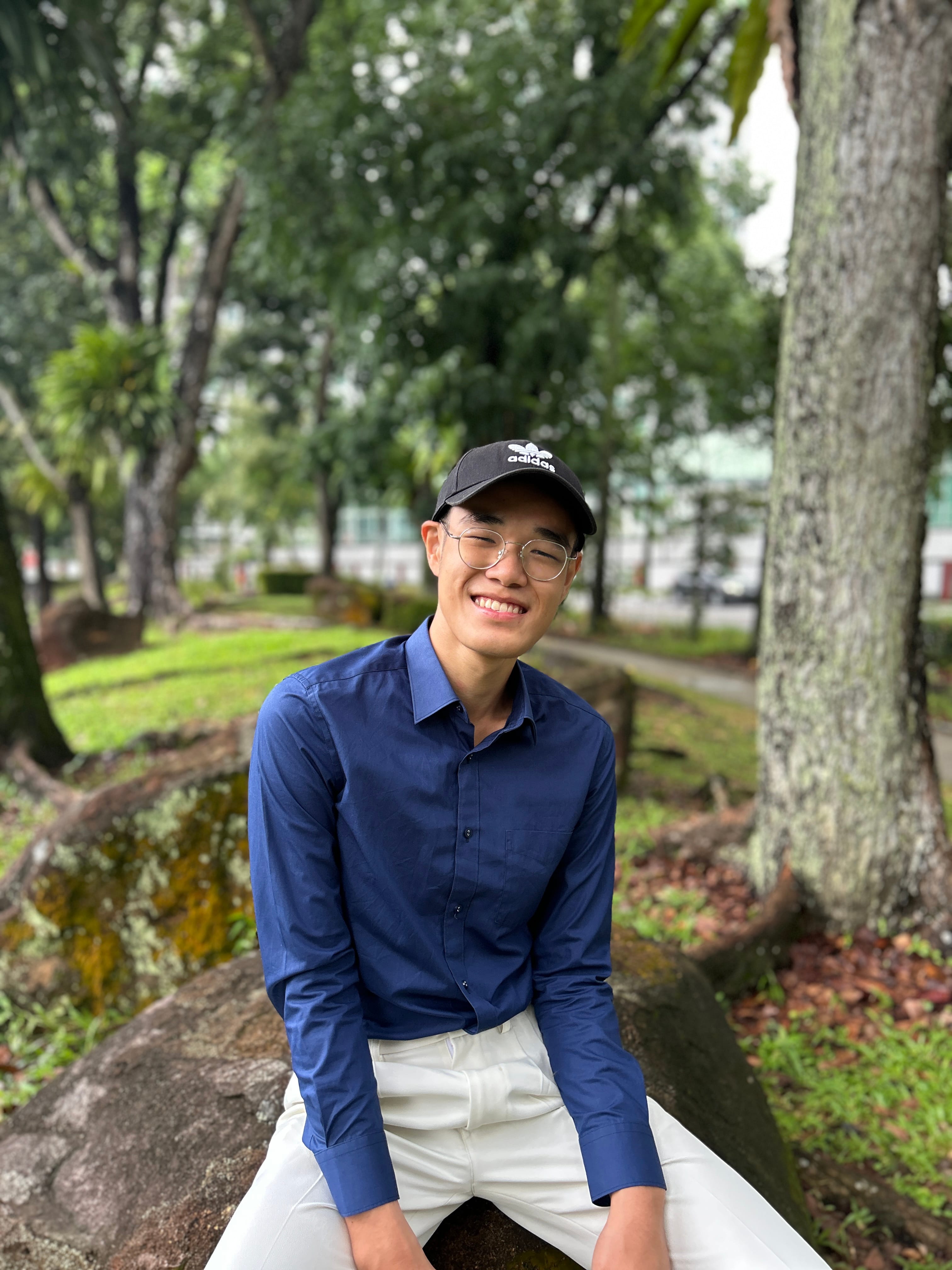 Young man smiling, wearing glasses, black Adidas cap, blue shirt, white pants, sitting on a rock in a green park.