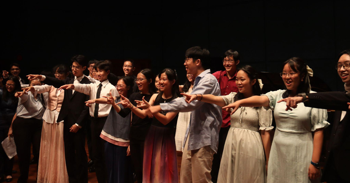A group of smiling young people on a dark stage, pointing forward and laughing together after a performance.