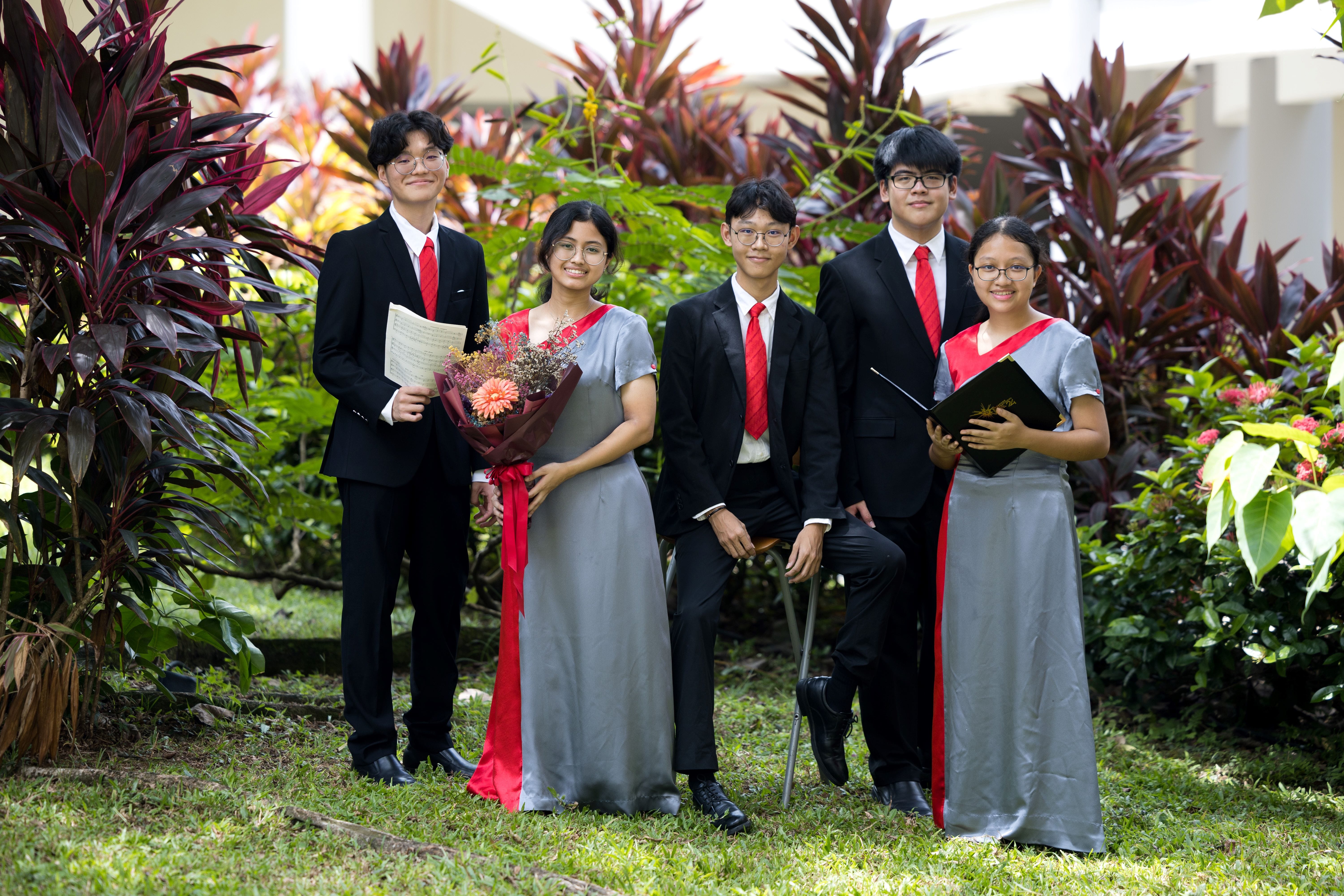 Five young students in formal black suits and gray dresses with red sashes pose outdoors among tropical plants.