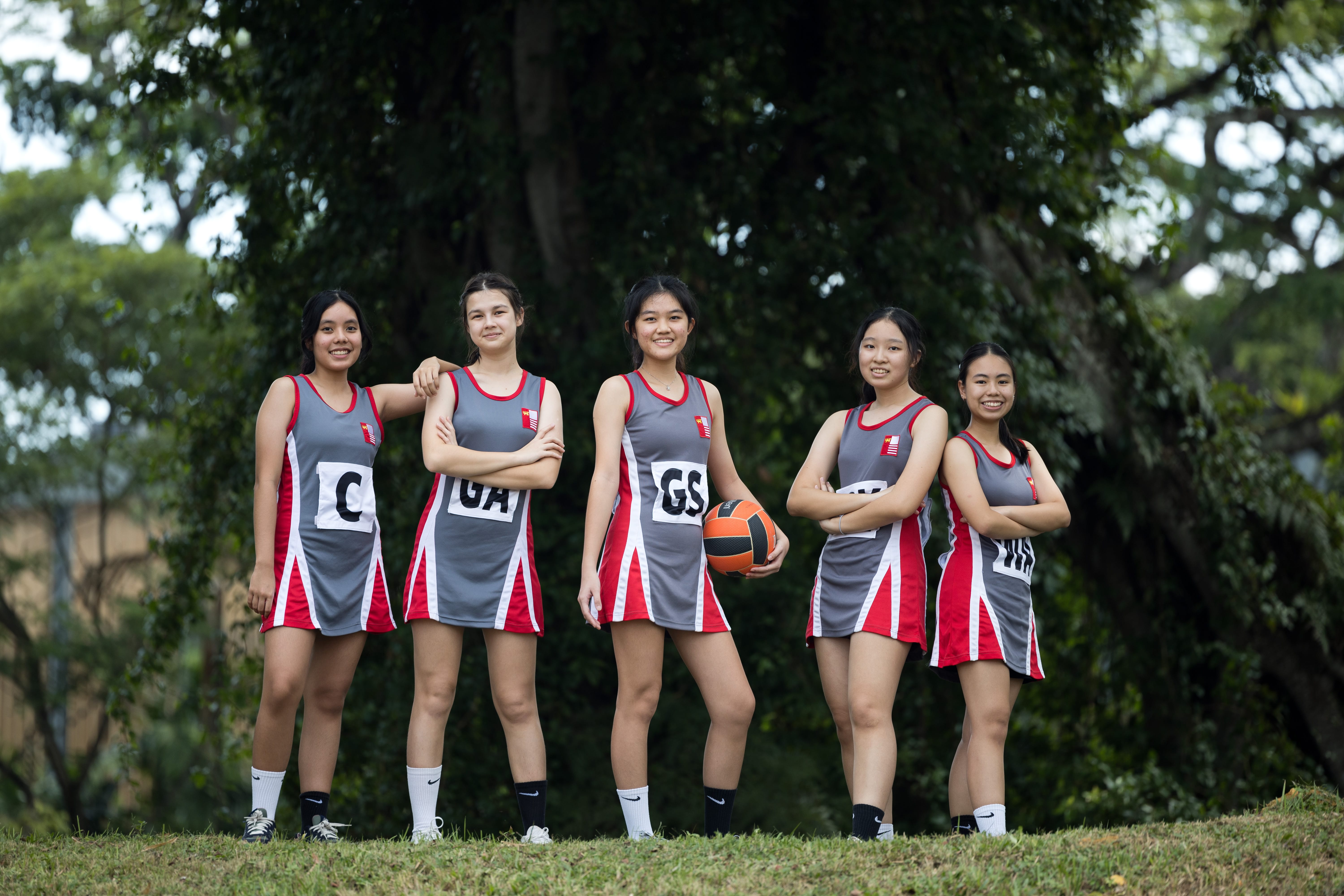Five girls in gray and red sports dresses pose on grass; one holds a basketball, trees behind.