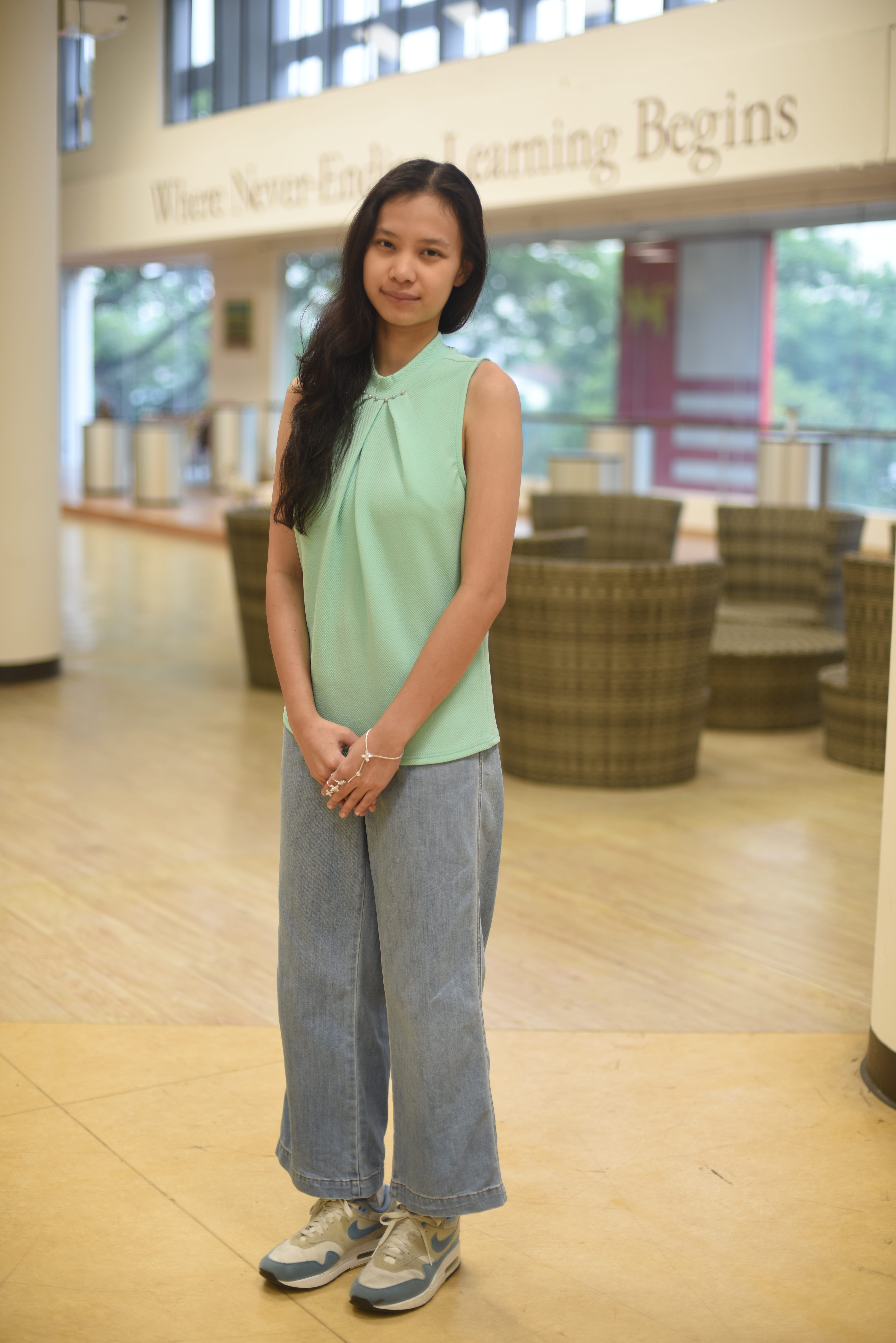 Person in mint sleeveless top and wide-leg jeans stands indoors near round chairs and tall windows.