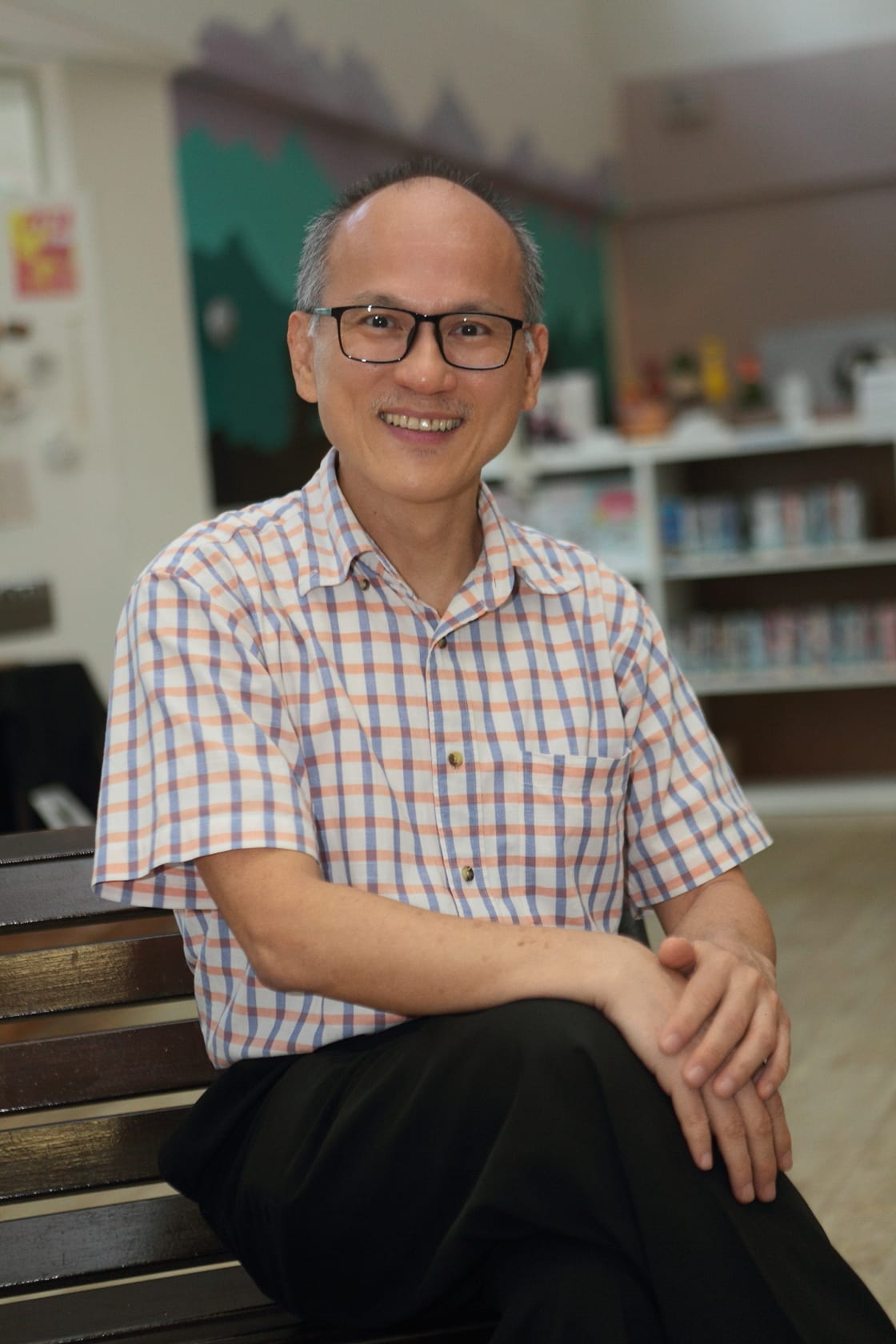 A smiling middle-aged man with glasses in a plaid shirt sits on a bench with bookshelves in the background.