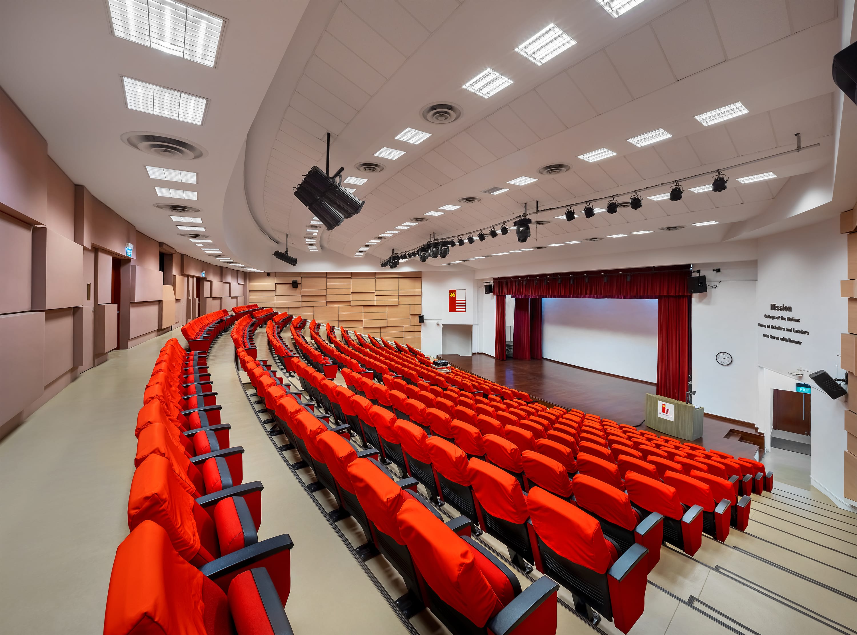 Empty lecture hall with curved rows of red seats facing a stage with red curtains and a white screen.