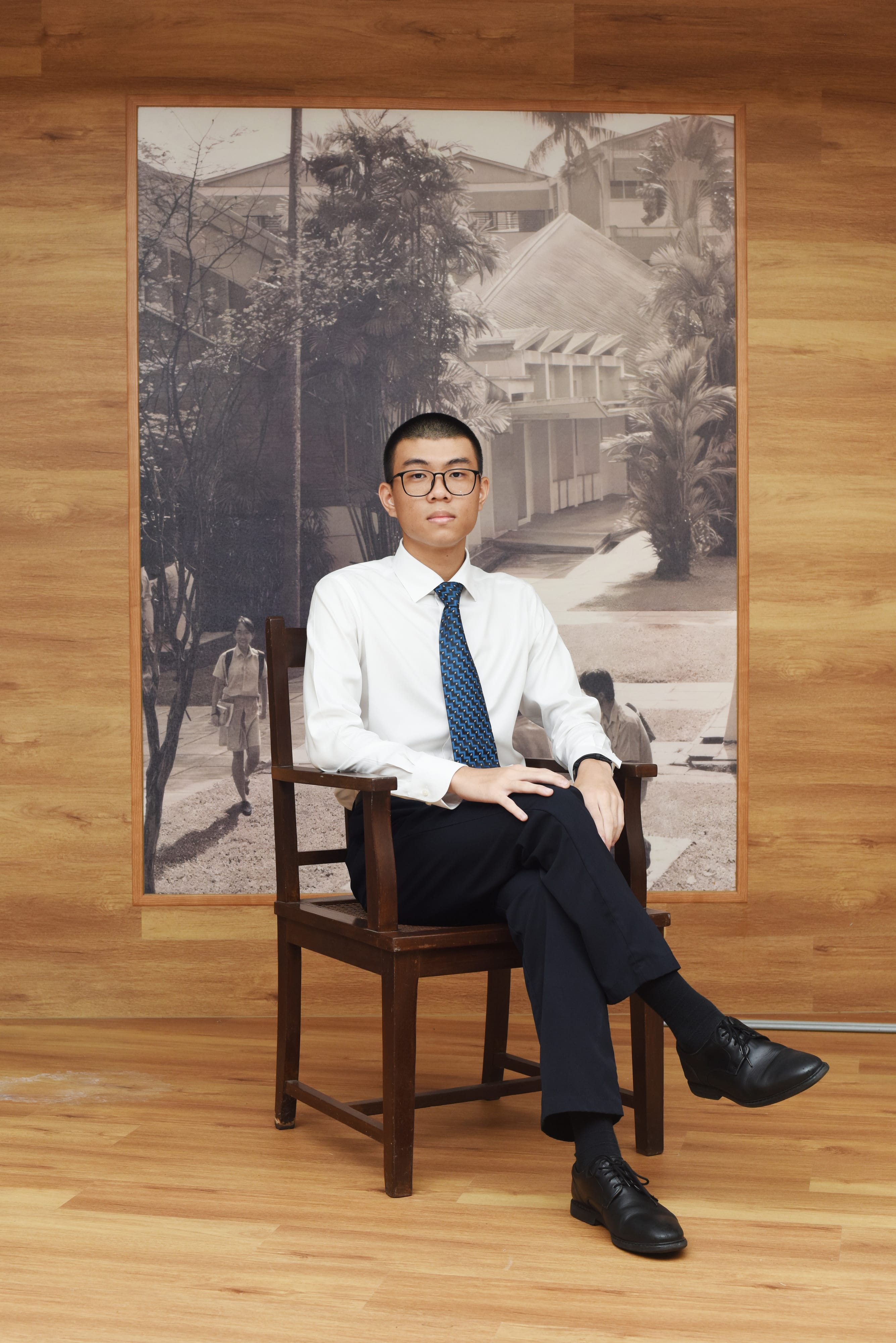 Young man in white shirt and blue tie sits cross-legged on a wooden chair before a large sepia school photograph.