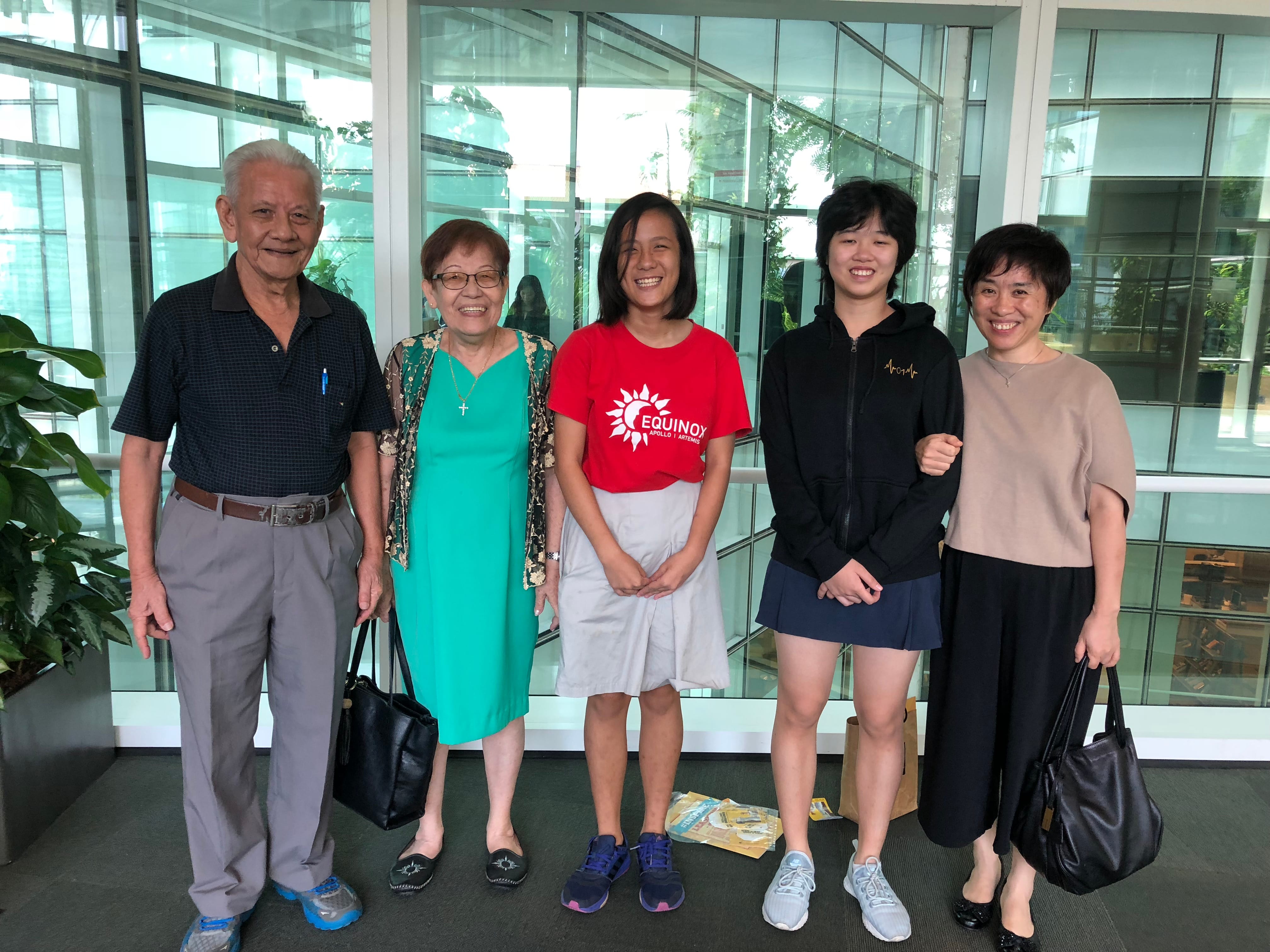 Five smiling people of various ages pose together in front of a glass building facade outdoors.