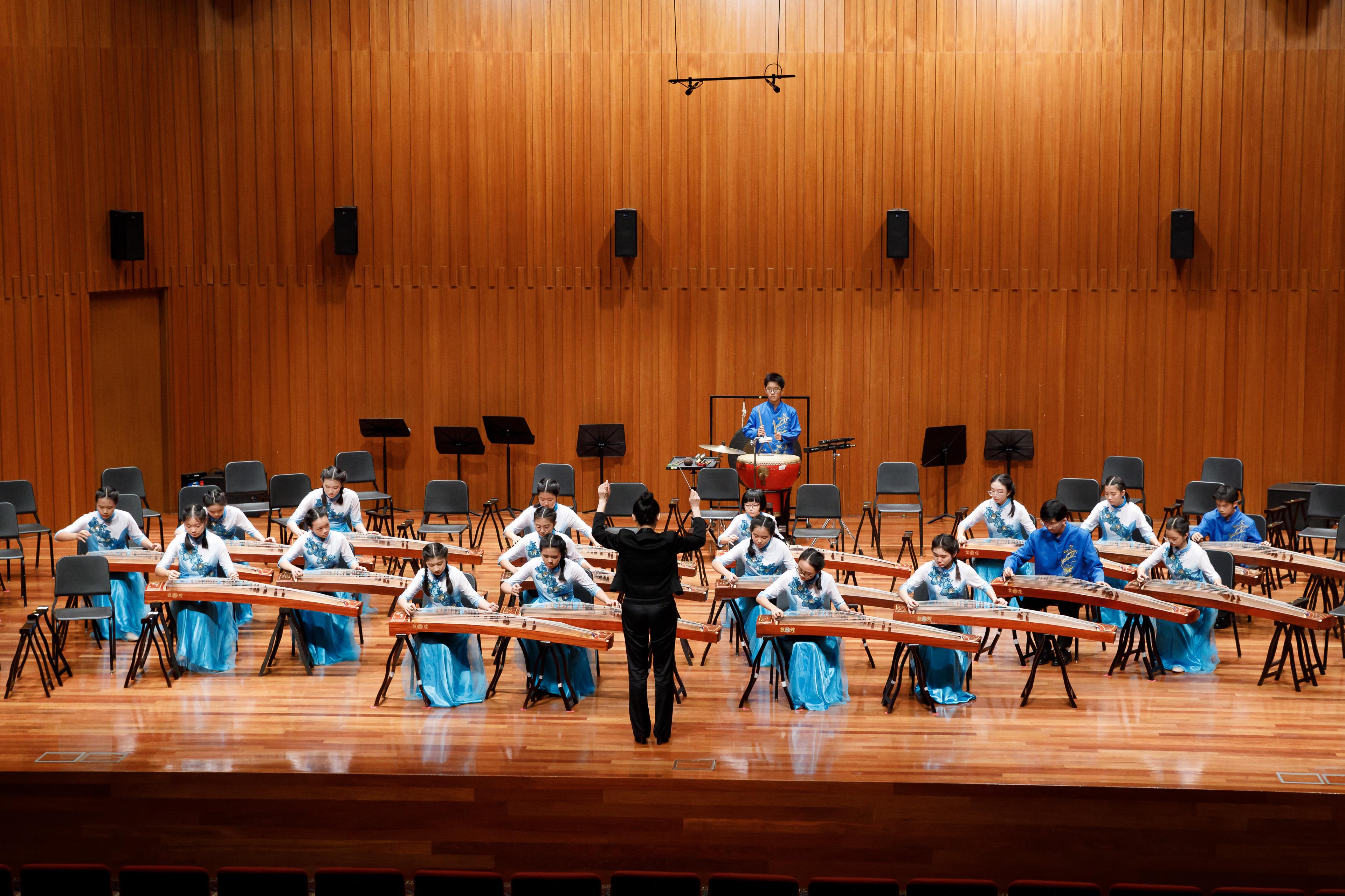 A conductor leads young musicians in blue dresses playing guzheng instruments on a wooden concert stage.