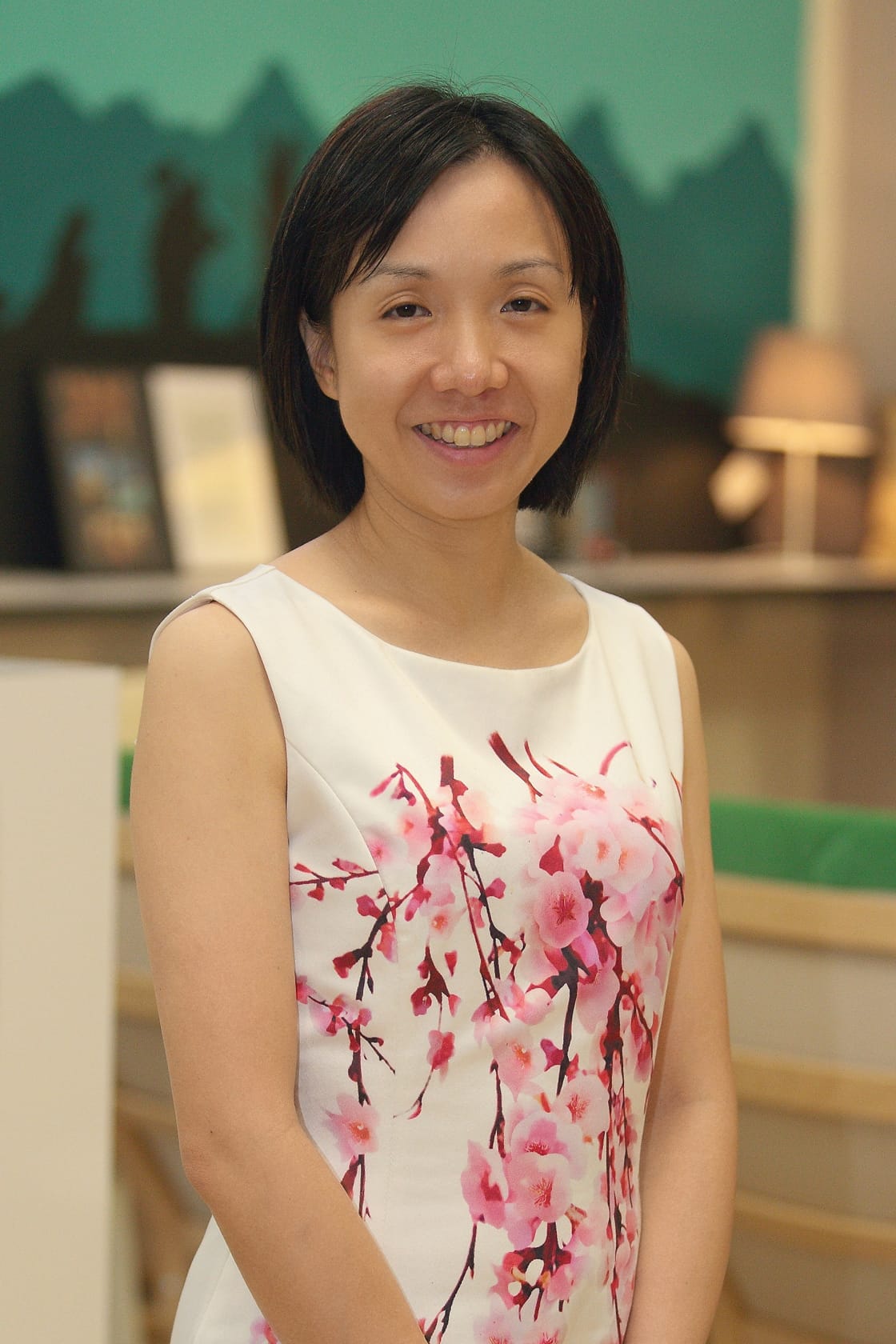 A smiling woman with short black hair wearing a white sleeveless dress with pink cherry blossom print.