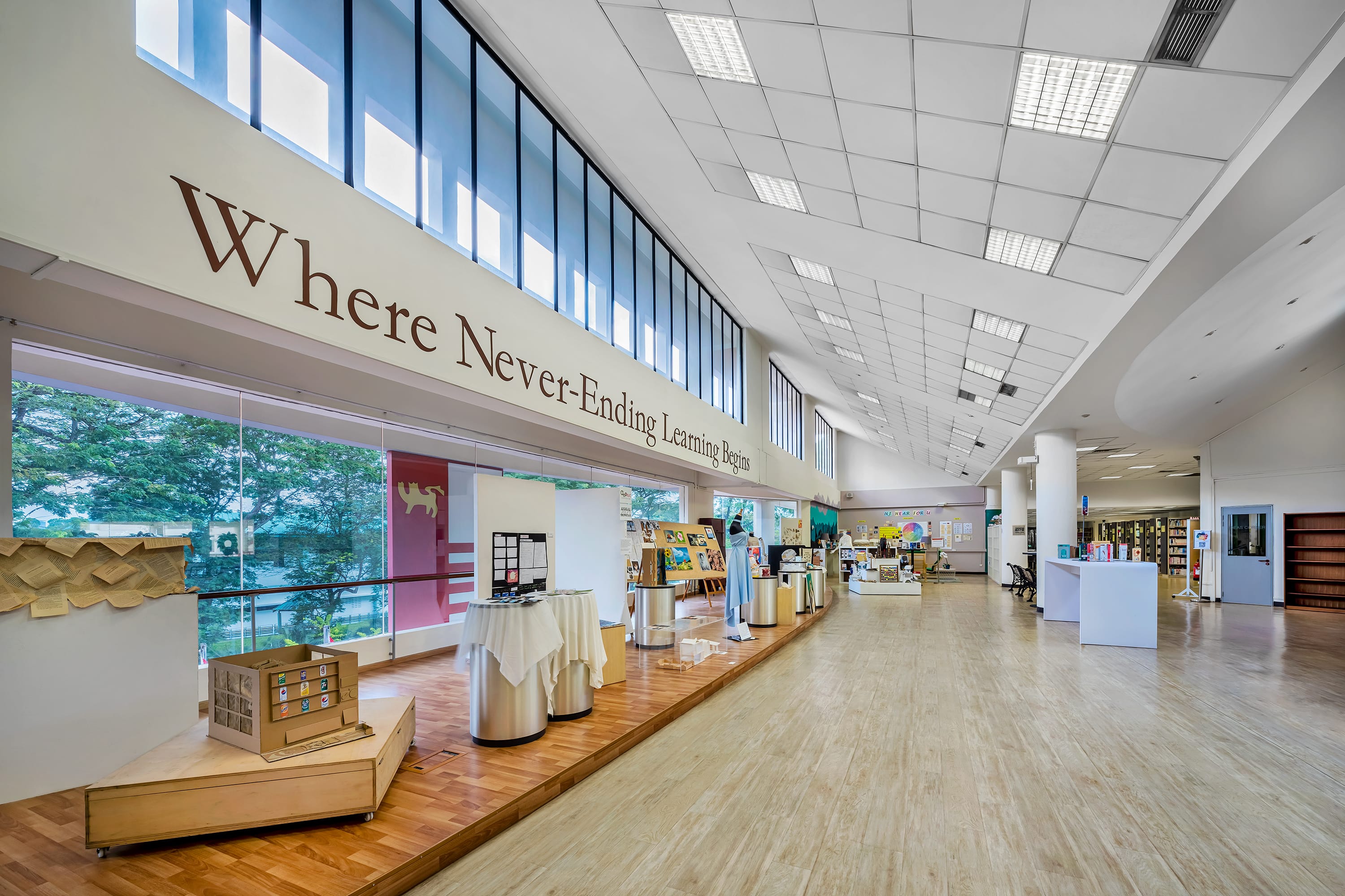 Spacious library hall with tall windows, display tables, and wall text reading Where Never-Ending Learning Begins