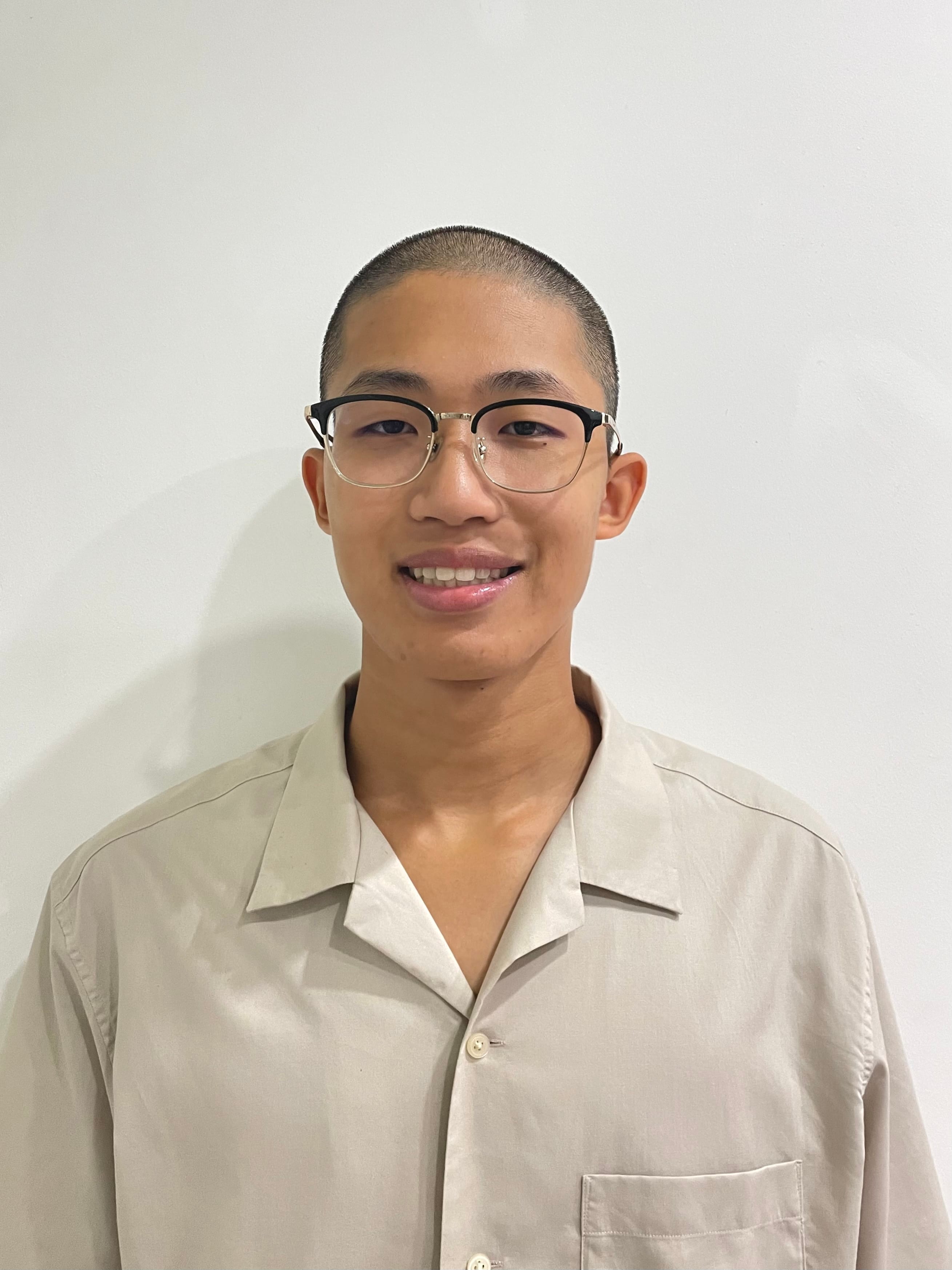 Young man with a buzz cut and black-rimmed glasses, smiling, wearing a beige collared shirt against a white wall.