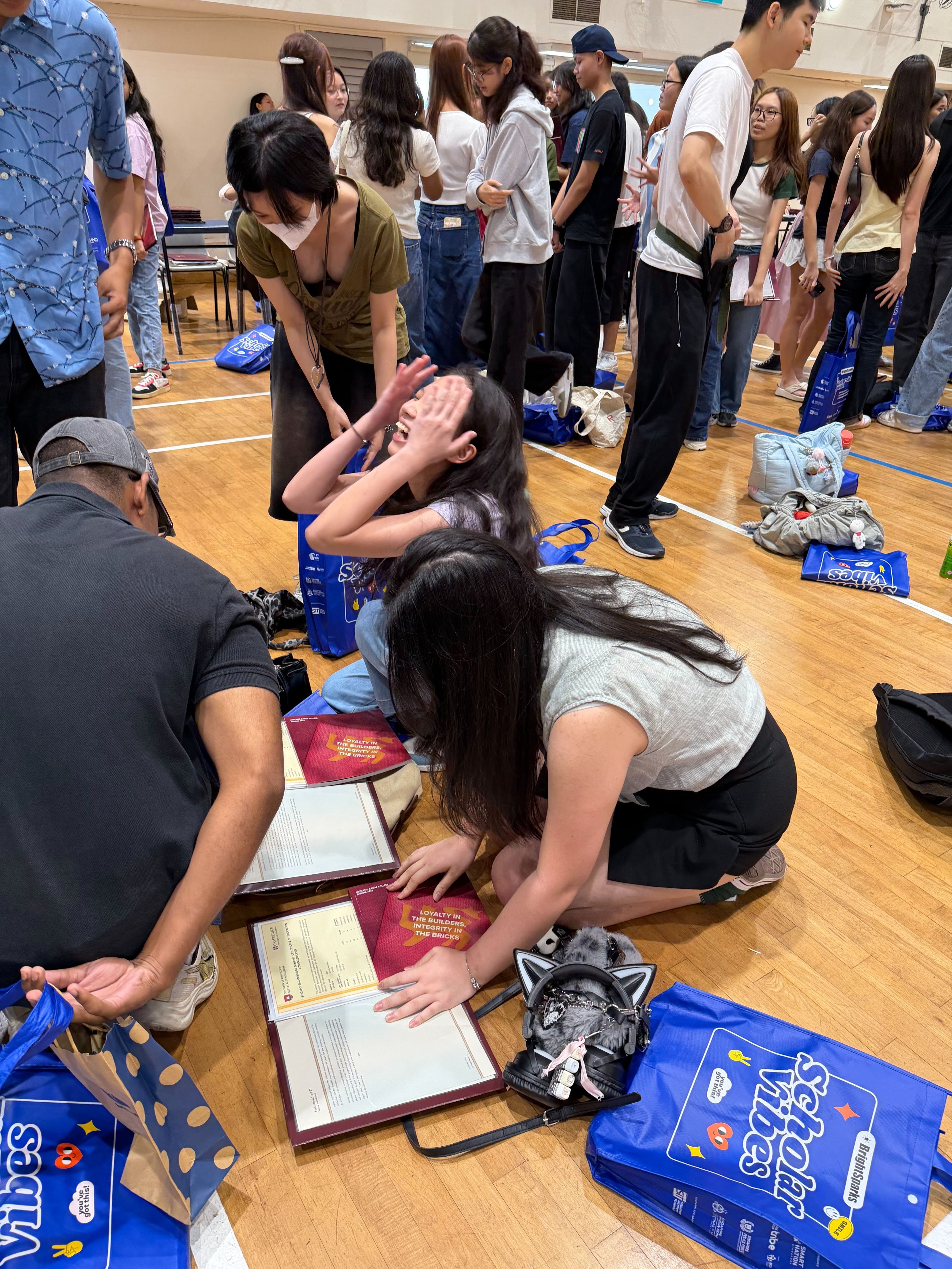 Students gather in a gymnasium, some kneeling on the floor sorting documents and blue Scholar Vibes tote bags.