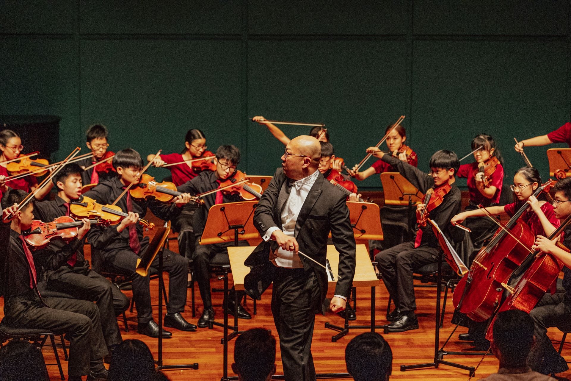 A bald conductor in a black suit leads a youth string orchestra dressed in red and black on a wooden stage.