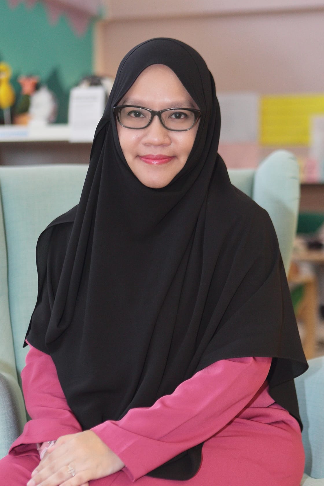 A smiling woman wearing a black hijab, glasses, and a pink top, seated in a light blue chair indoors.