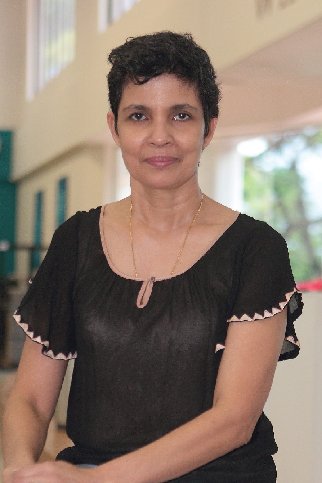 A woman with short dark hair wearing a black blouse and gold necklace stands indoors with a blurred background.