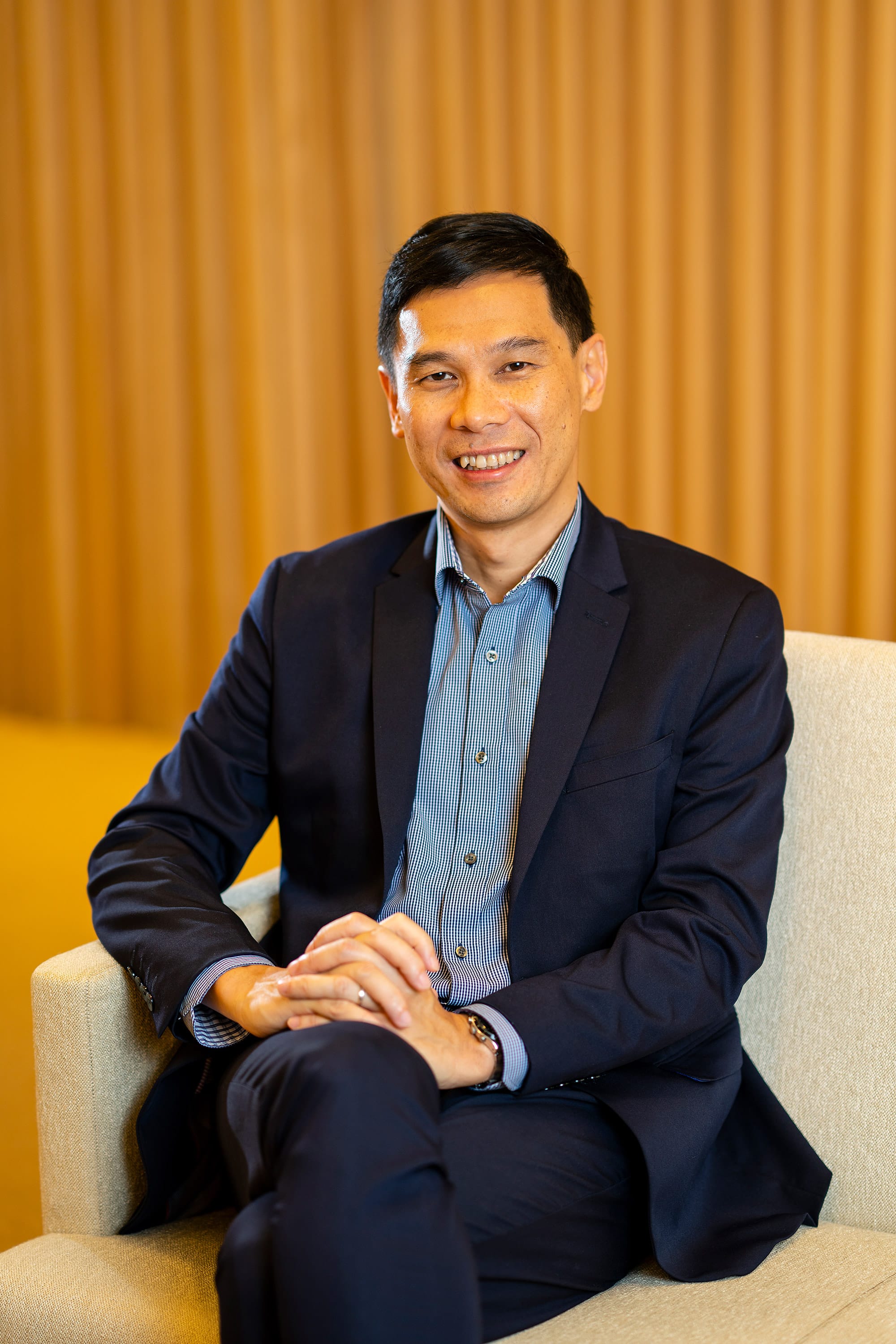A smiling man in a navy suit and blue shirt sits in a light chair with hands clasped, before a warm wooden wall.