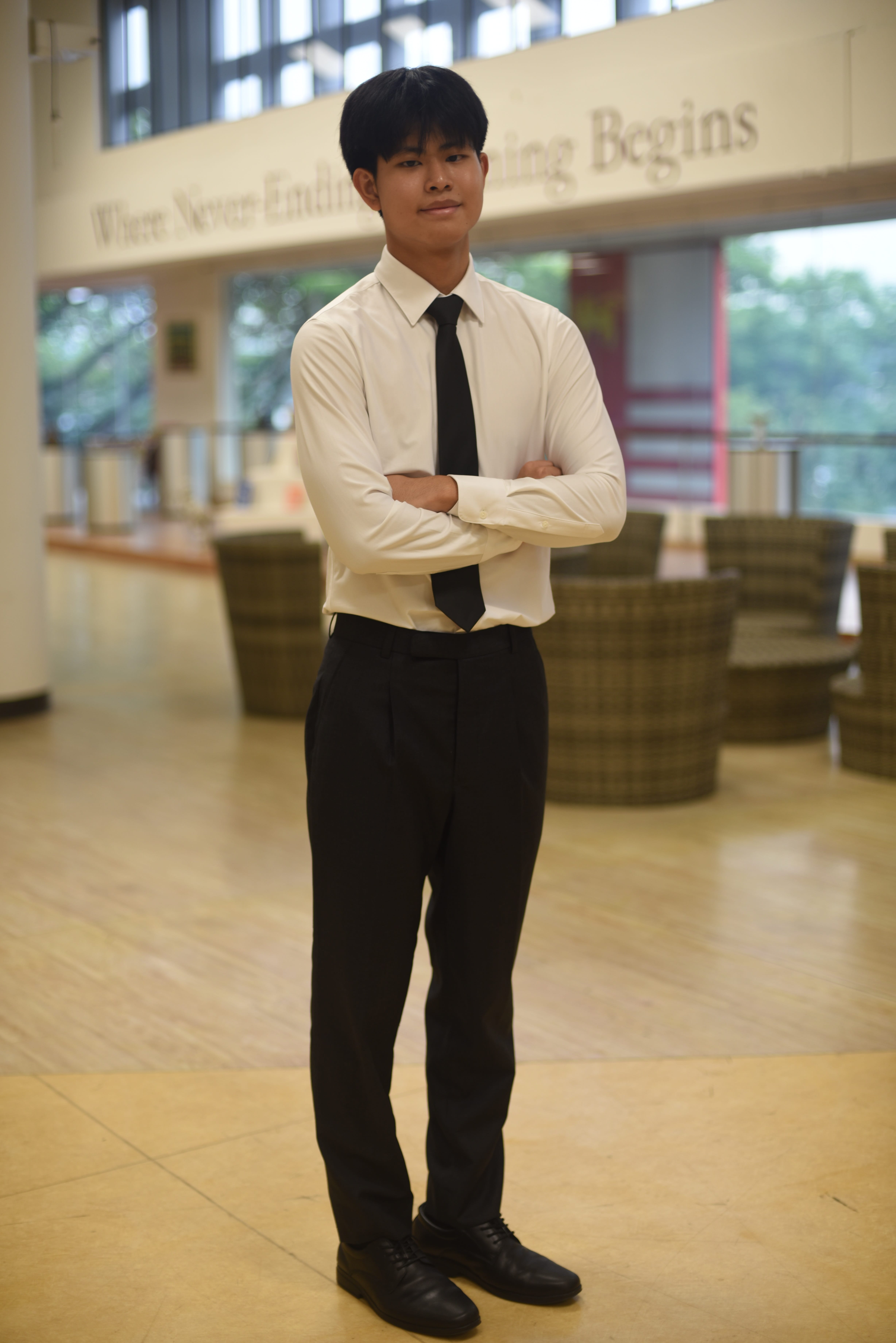 Person in white shirt and black tie stands with arms crossed in a bright lobby with chairs behind.