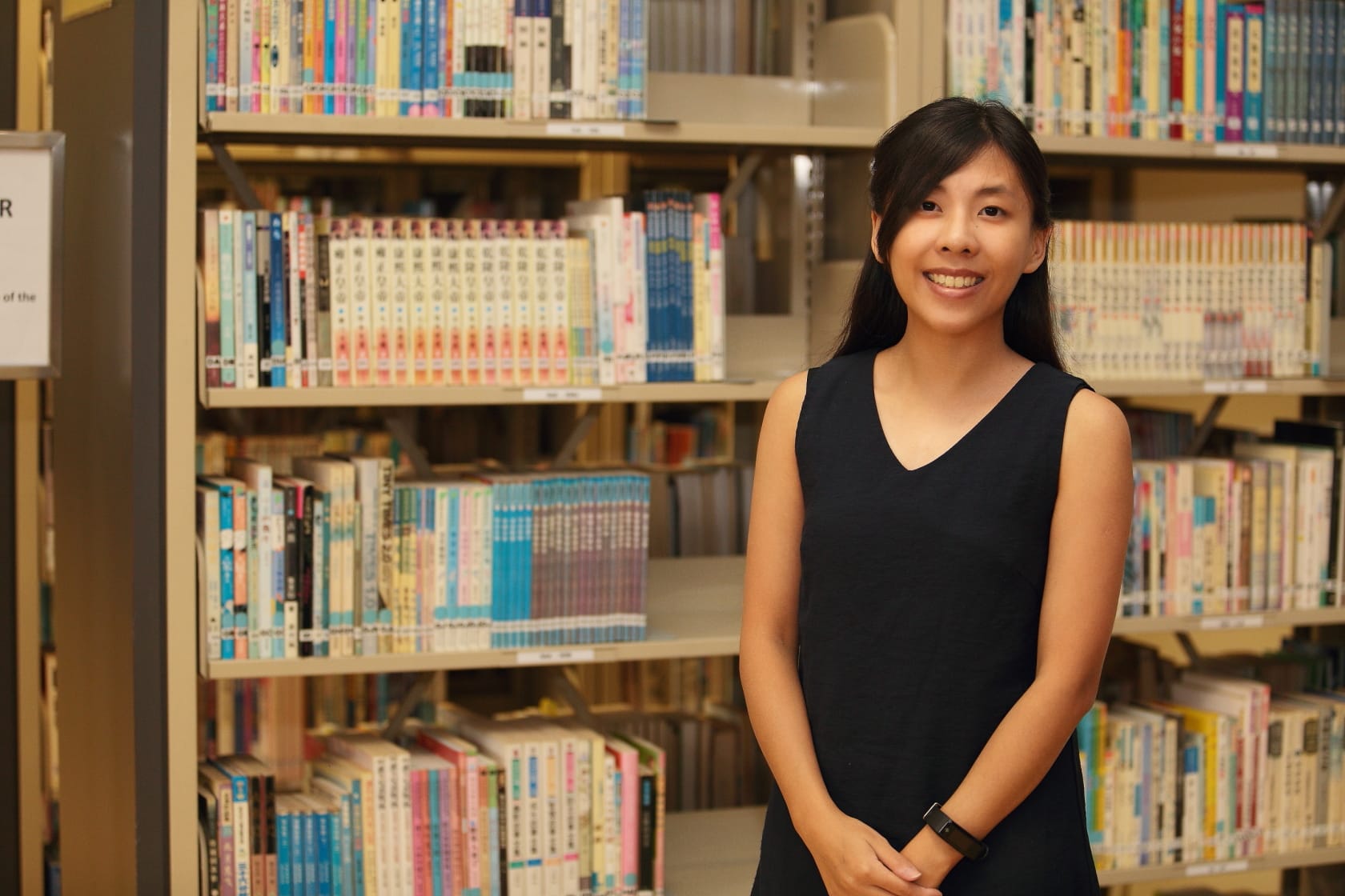 A smiling young woman in a black sleeveless dress stands in front of shelves filled with colorful books in a library.