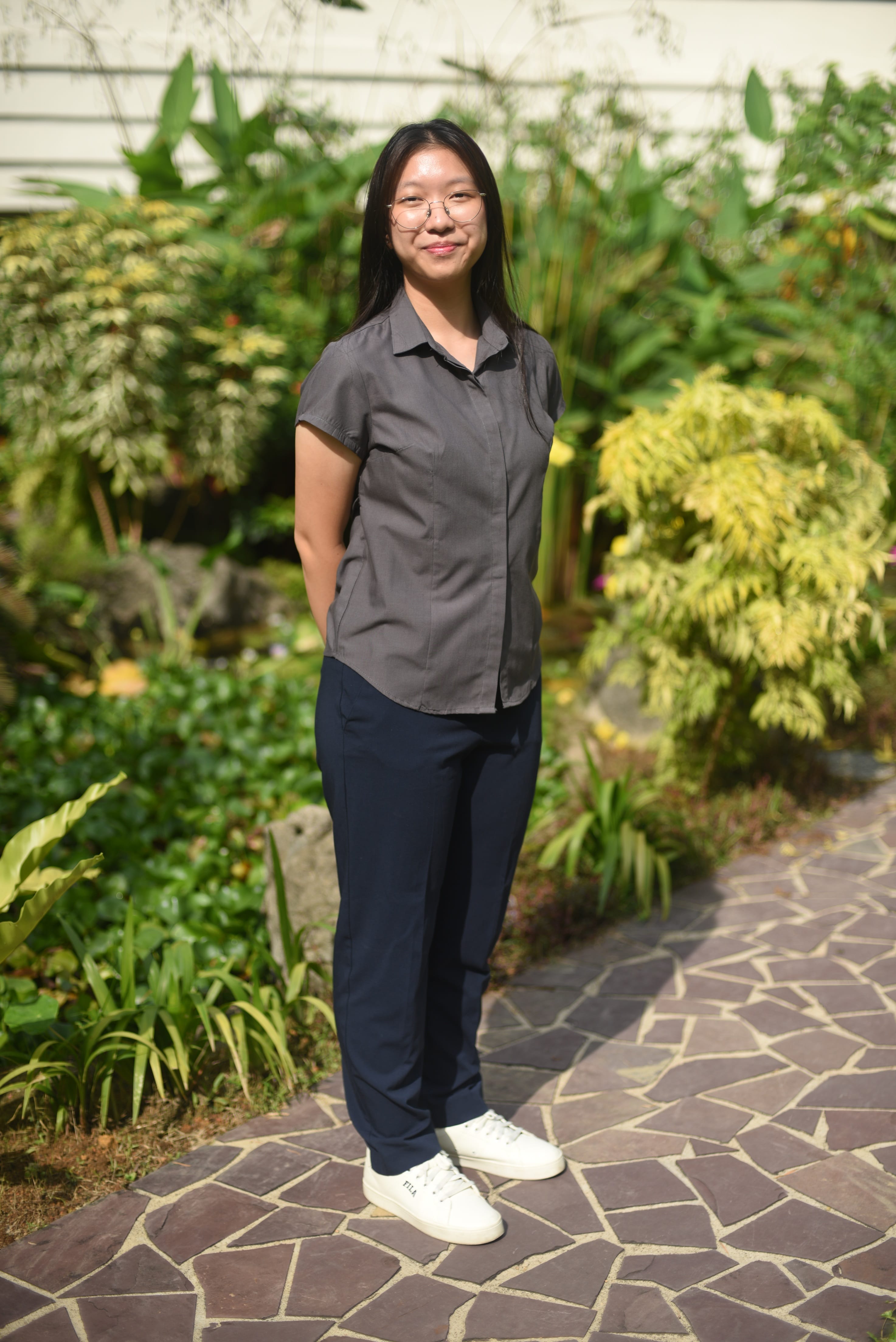 Young woman with glasses wearing a gray shirt and navy pants stands smiling on a stone path surrounded by green plants.