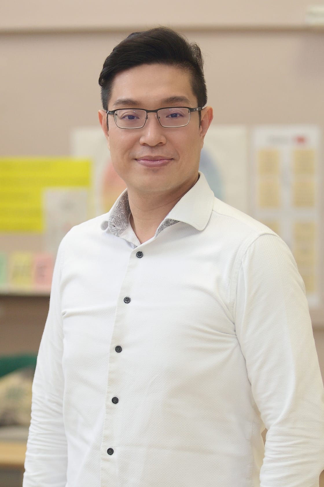 A man with glasses and dark hair wearing a white button-up shirt, smiling in front of a blurred indoor background.