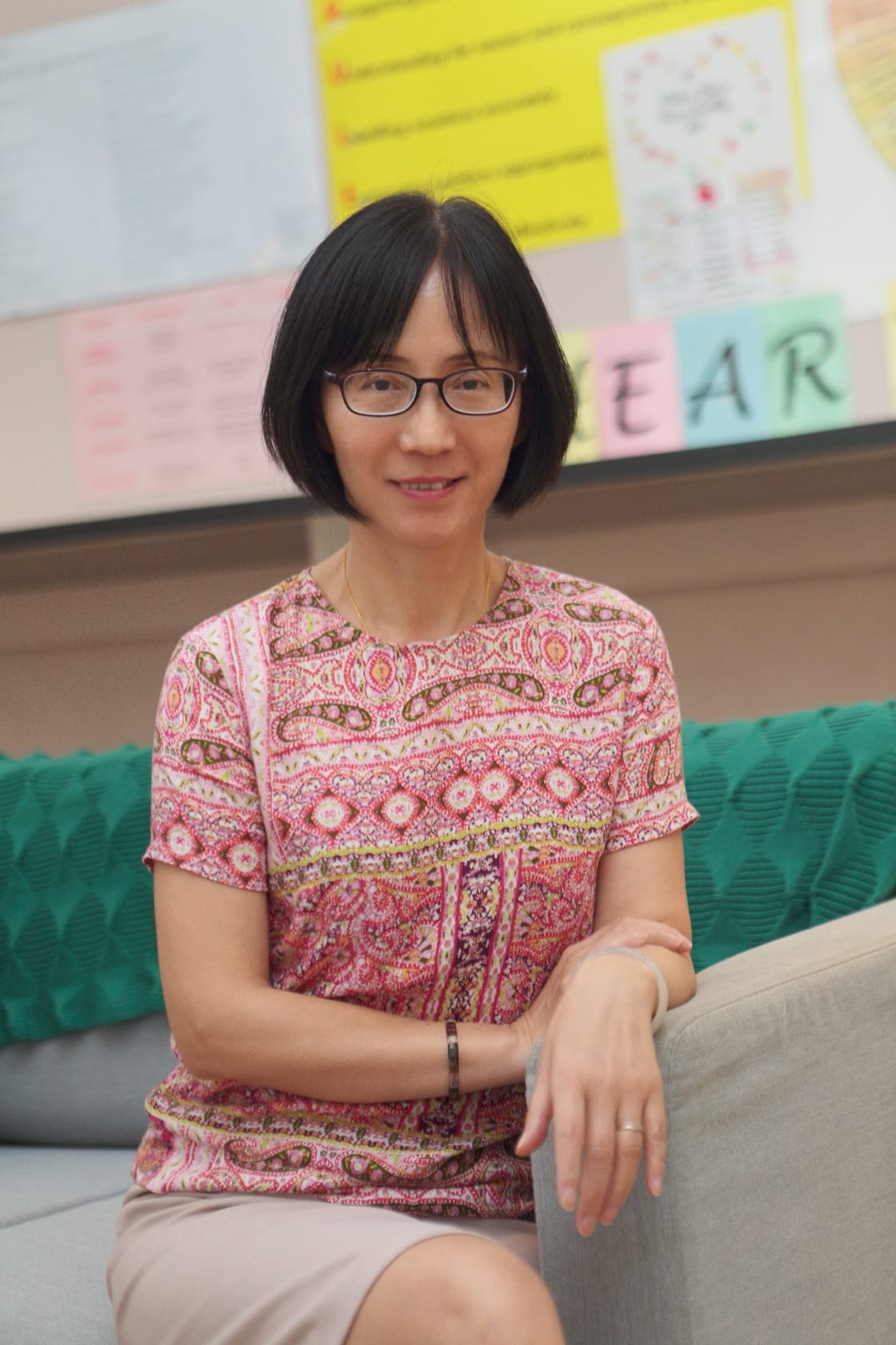 Woman with short black hair and glasses wearing a pink patterned top, seated on a gray sofa with teal cushions.